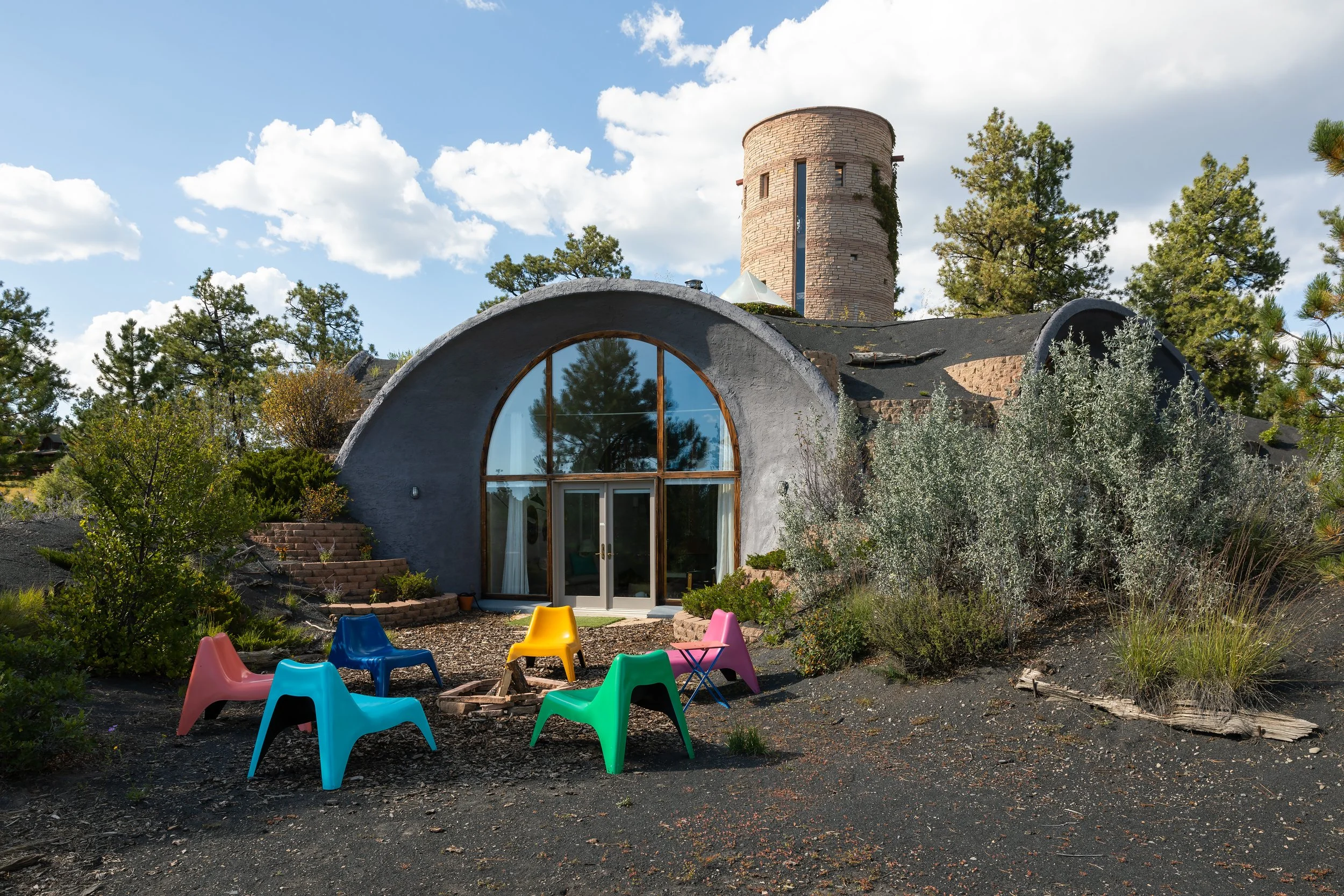 Unique house with a rounded arched window, stone chimney, and colorful outdoor chairs around a fire pit amid trees and shrubs.