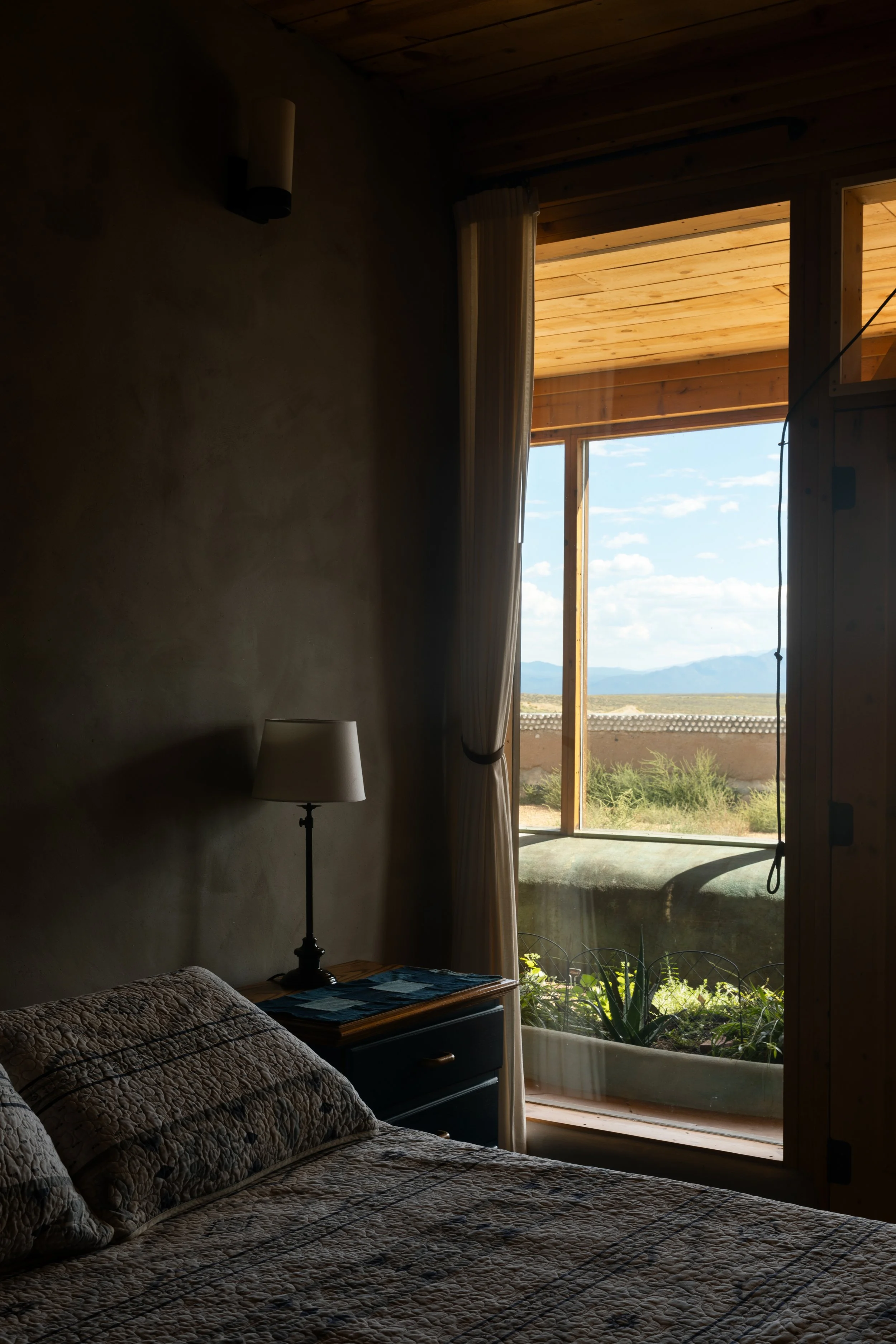 A cozy bedroom with a bed, patterned quilt, and a pillow, illuminated by sunlight coming through a large open window with a view of landscape and mountains in the distance.