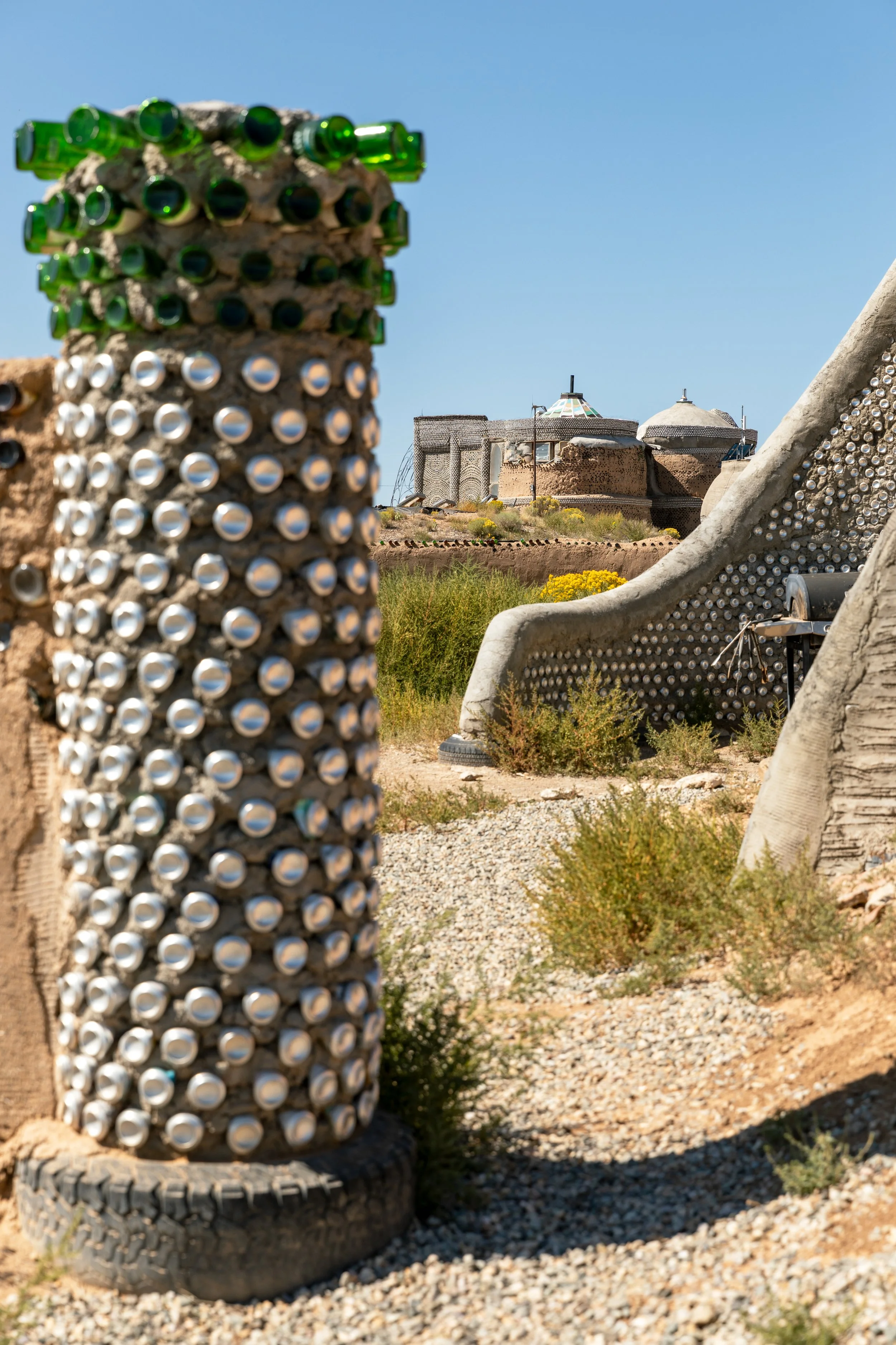 Outdoor art installation with vertical columns made of glass bottles and a concrete slide covered in glass bottles, set in a desert landscape with buildings in the background.