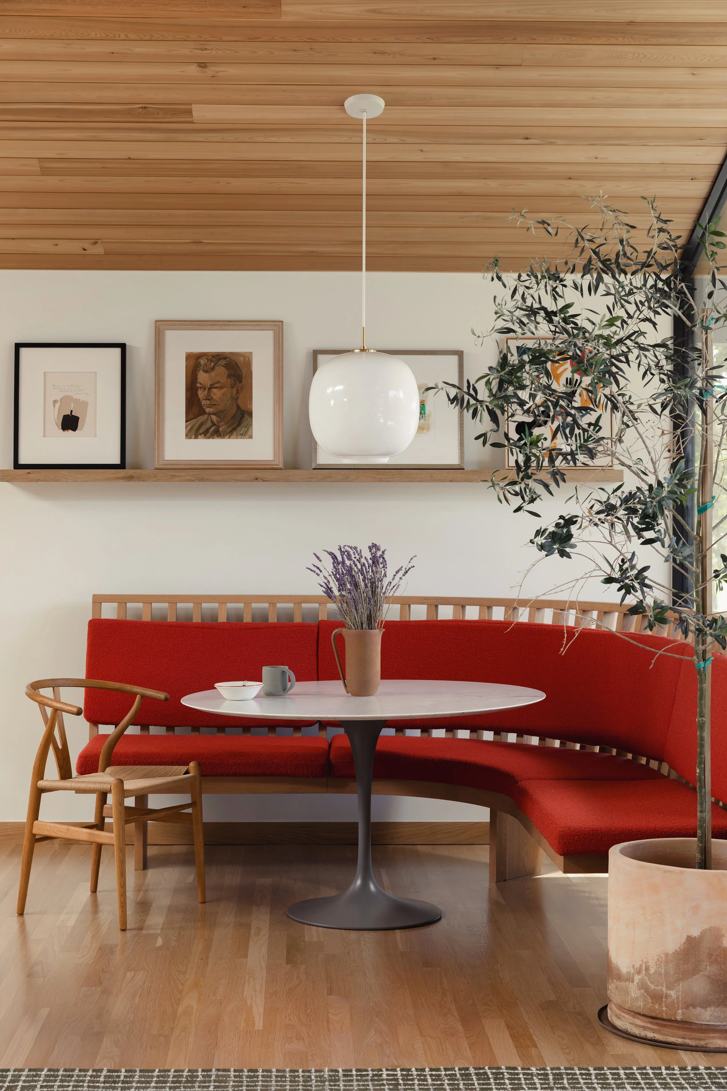 Living room with a round table, red sofa, wooden chair, potted lamp, wall art, and a hanging light fixture.