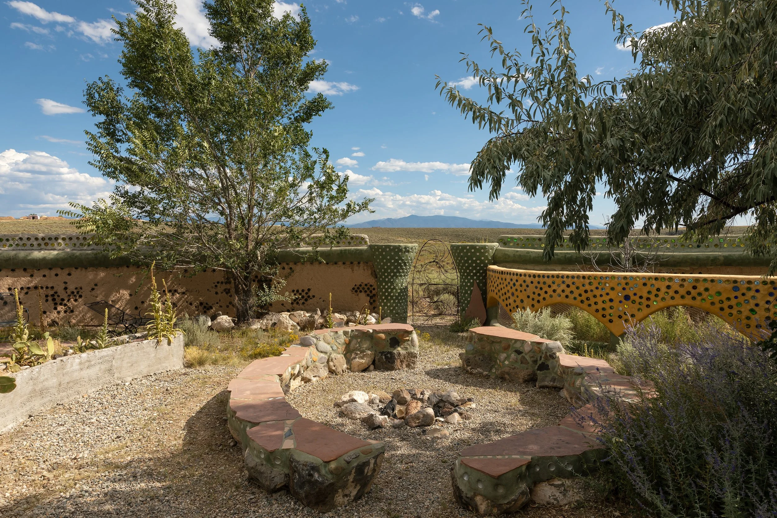 Desert garden with two colorful concrete benches, a fire pit made of rocks, and a low concrete wall with a metal gate. Two trees provide shade under a blue sky with clouds, and mountains are visible in the distance.