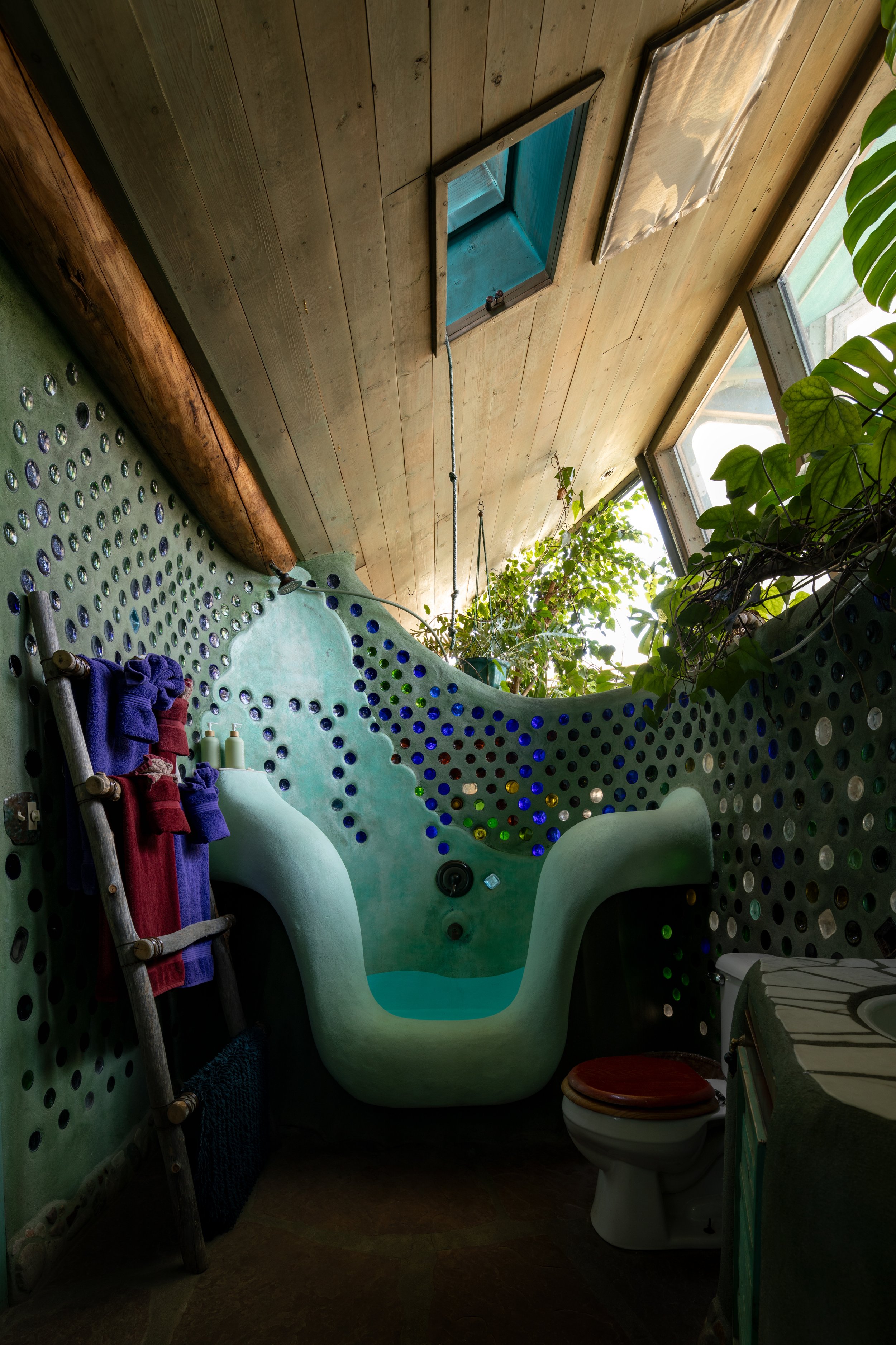 Eclectic bathroom with a green custom bathtub, a wooden ladder with hanging towels, a toilet with a red seat cover, and windows with natural light, surrounded by greenery.