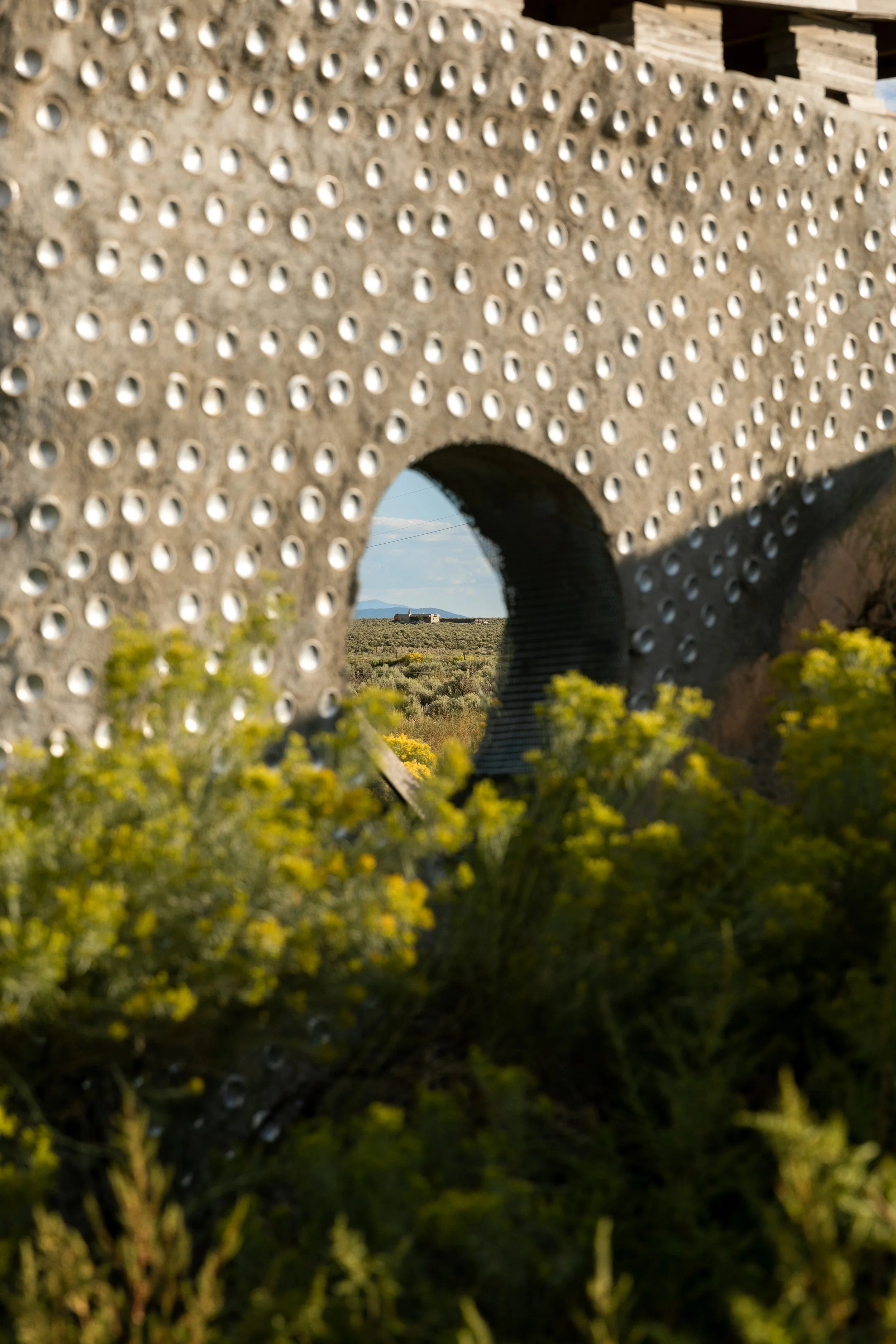 View through a circular opening in a perforated concrete wall, with yellow wildflowers in the foreground and a distant landscape with a building on a hill under a clear blue sky.