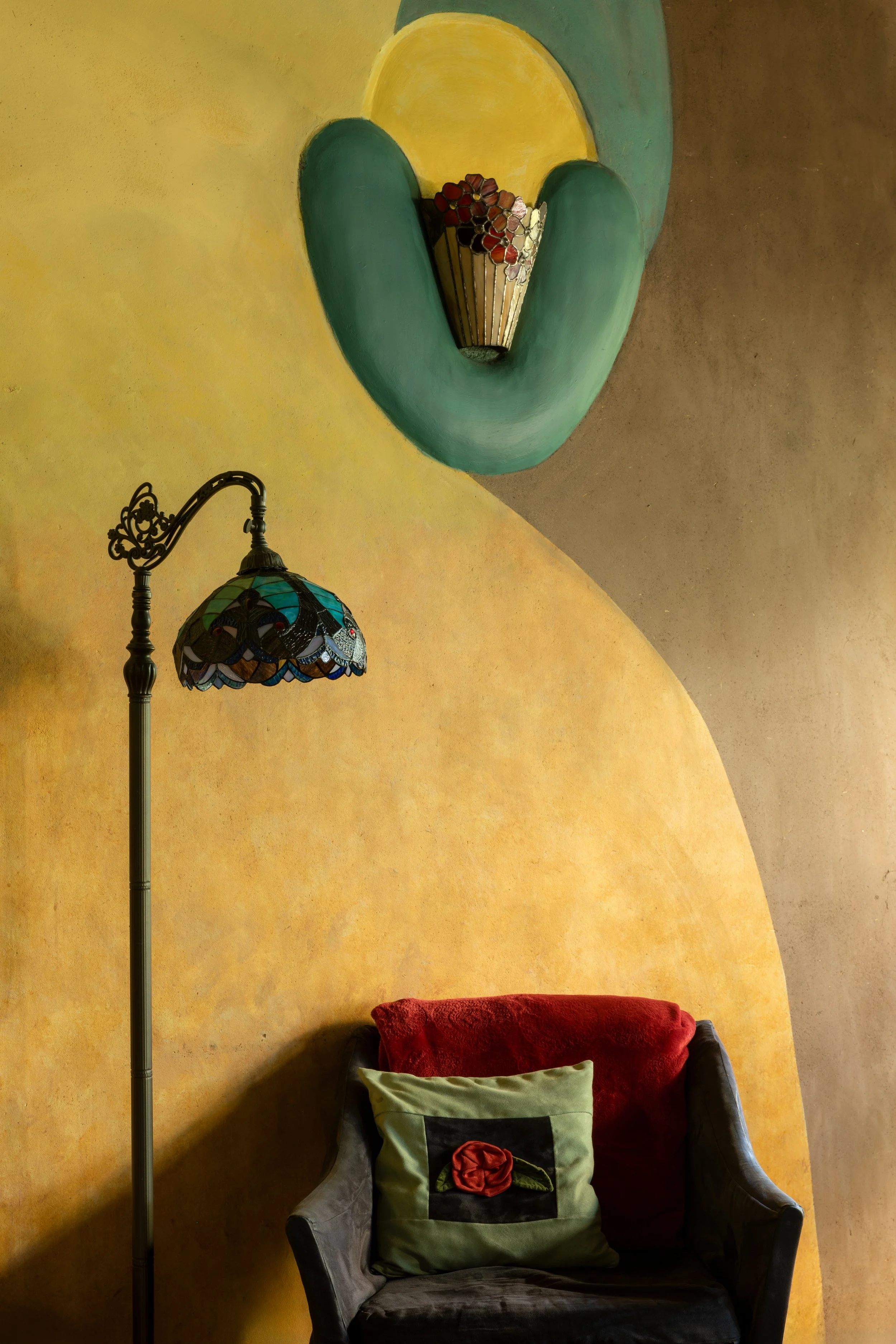 A corner of a room with a yellow and brown wall featuring a colorful mural. There is a black armchair with cushions, one green with a red flower and leaf embroidery, and a vintage-style standing lamp with a stained-glass shade.