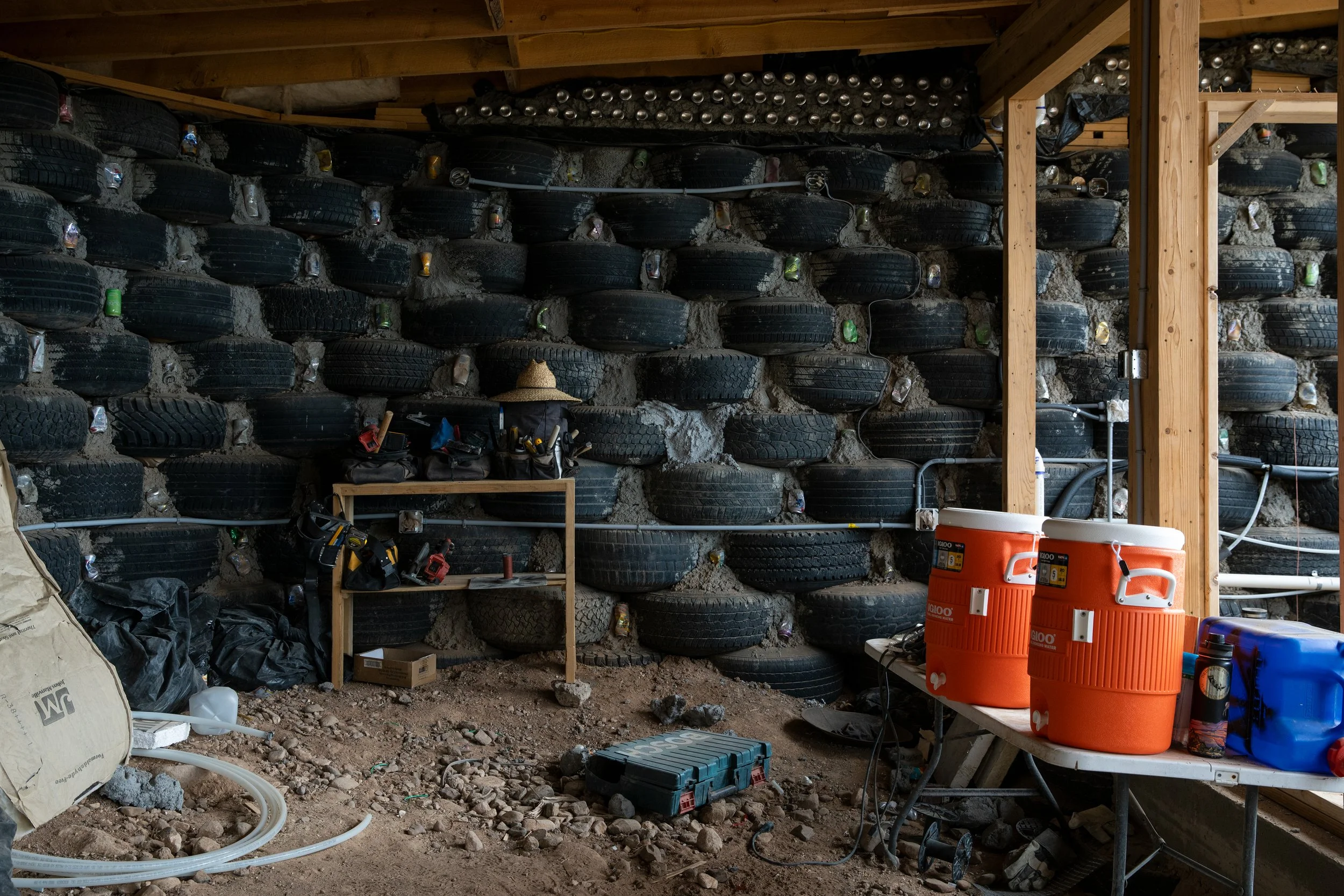 A storage area under construction with tires stacked against the wall, tools on a small table, and large orange coolers on a folding table, with construction materials and dirt on the ground.