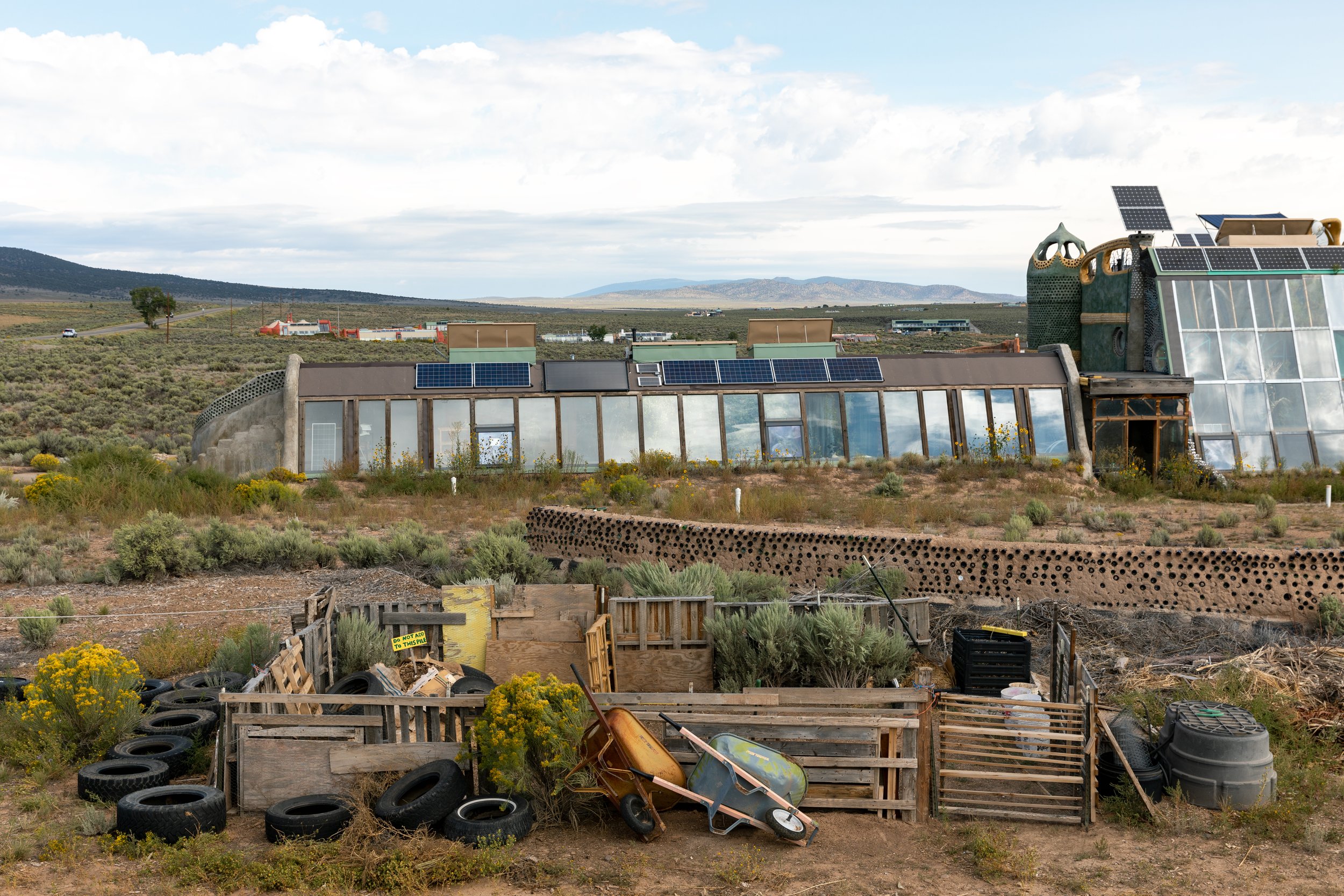A futuristic building with solar panels on the roof, located in a desert landscape. The building has large glass windows and a unique, artistic design with colorful pipes or structures on top. In the foreground, there is a pile of discarded tires, wo