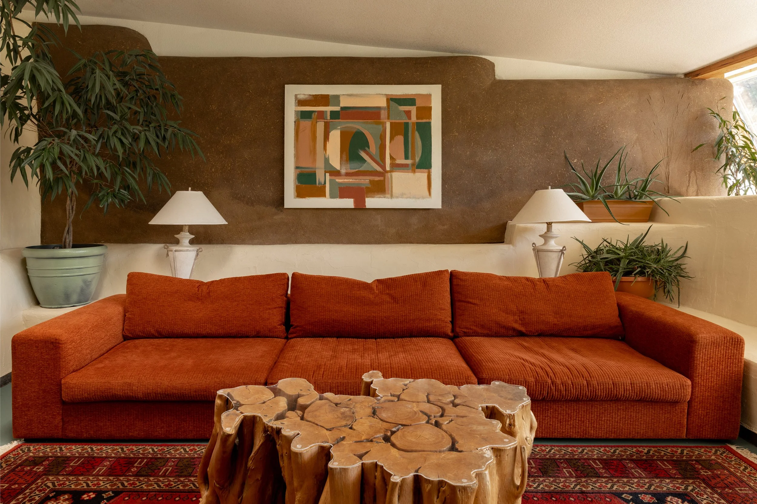 A living room with a burnt orange sofa, two white lamps, two potted plants, a wooden coffee table, a colorful abstract picture, and a patterned rug.