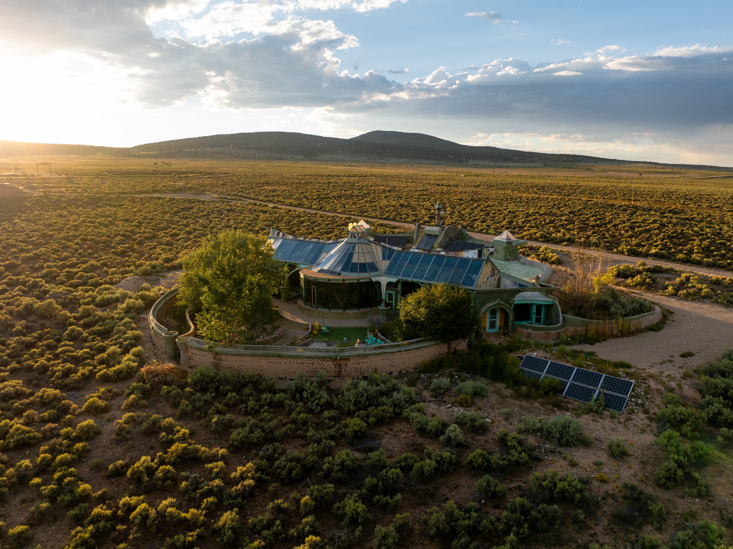 An aerial view of a house with a modern, eclectic architectural style, solar panels, and large glass windows, situated in a vast, open desert landscape during sunset.