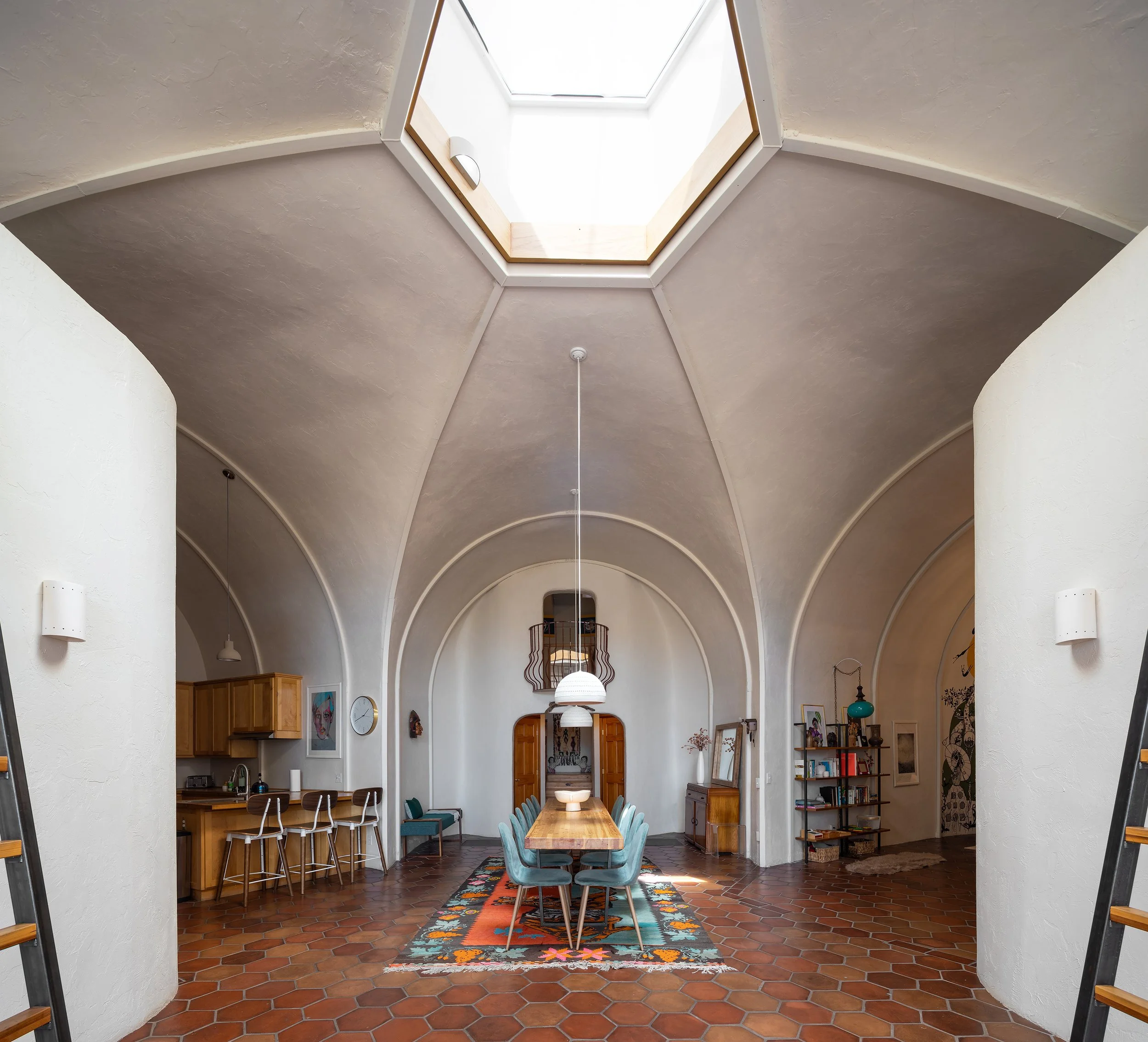 Interior view of a high-ceilinged dining room with a hexagonal skylight, a long wooden dining table with teal chairs, and a patterned area rug on terracotta tile flooring.