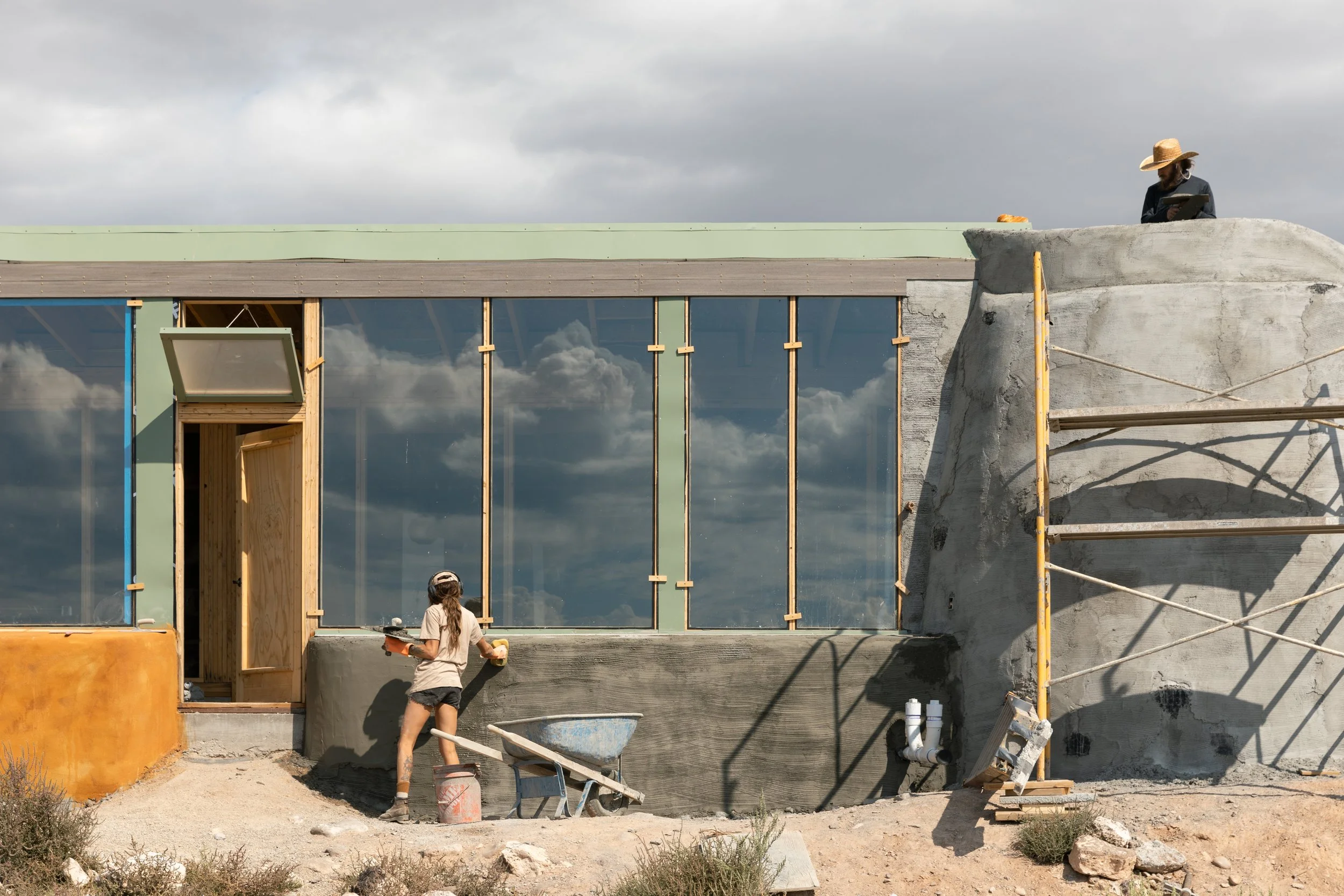 Construction workers building a modern house with large glass windows and a concrete and wood structure under a cloudy sky.