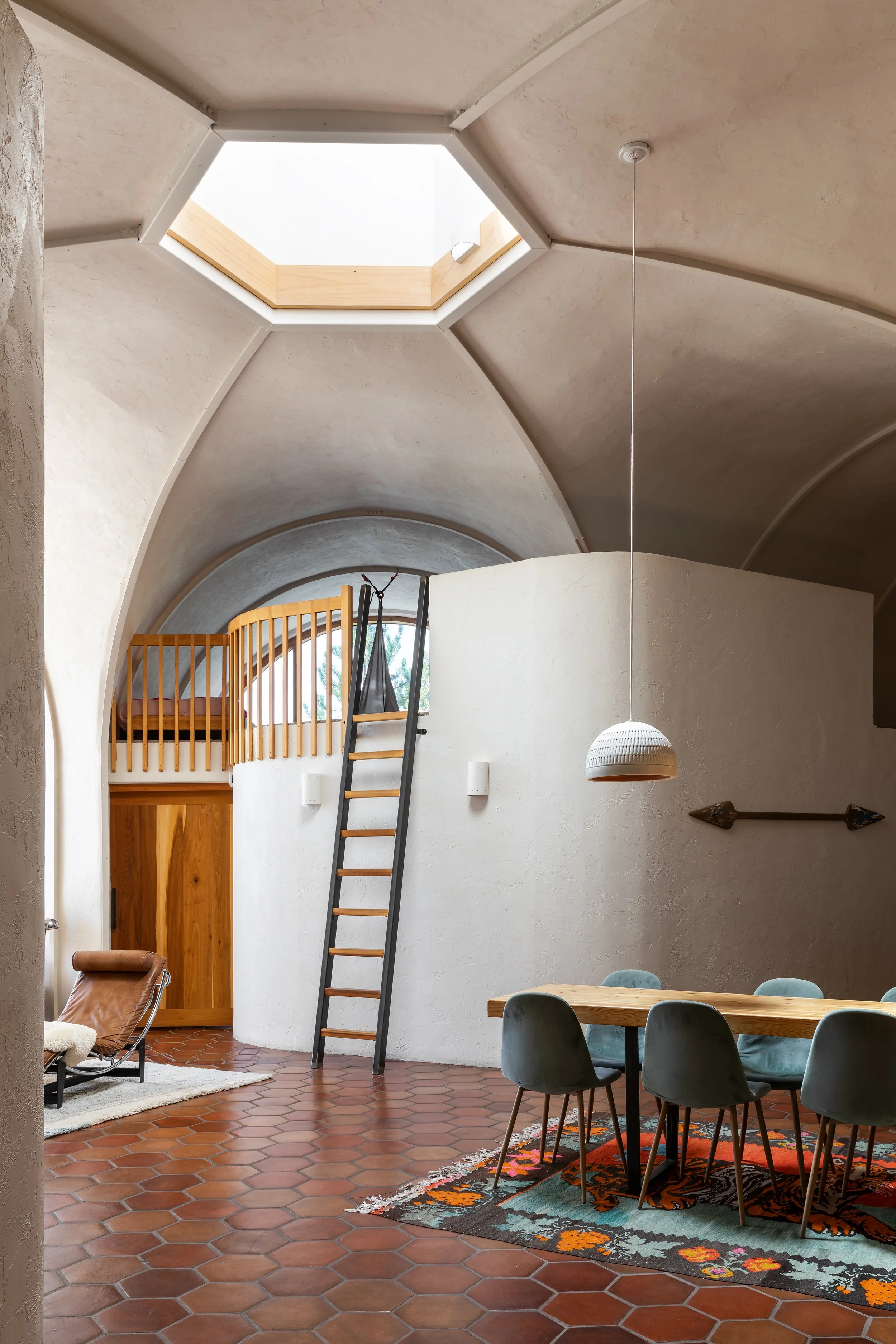 Interior of a modern home with a rounded ceiling, a skylight, a loft area with a wooden railing, a ladder, a dining table with chairs, and a rug on a terracotta tiled floor.
