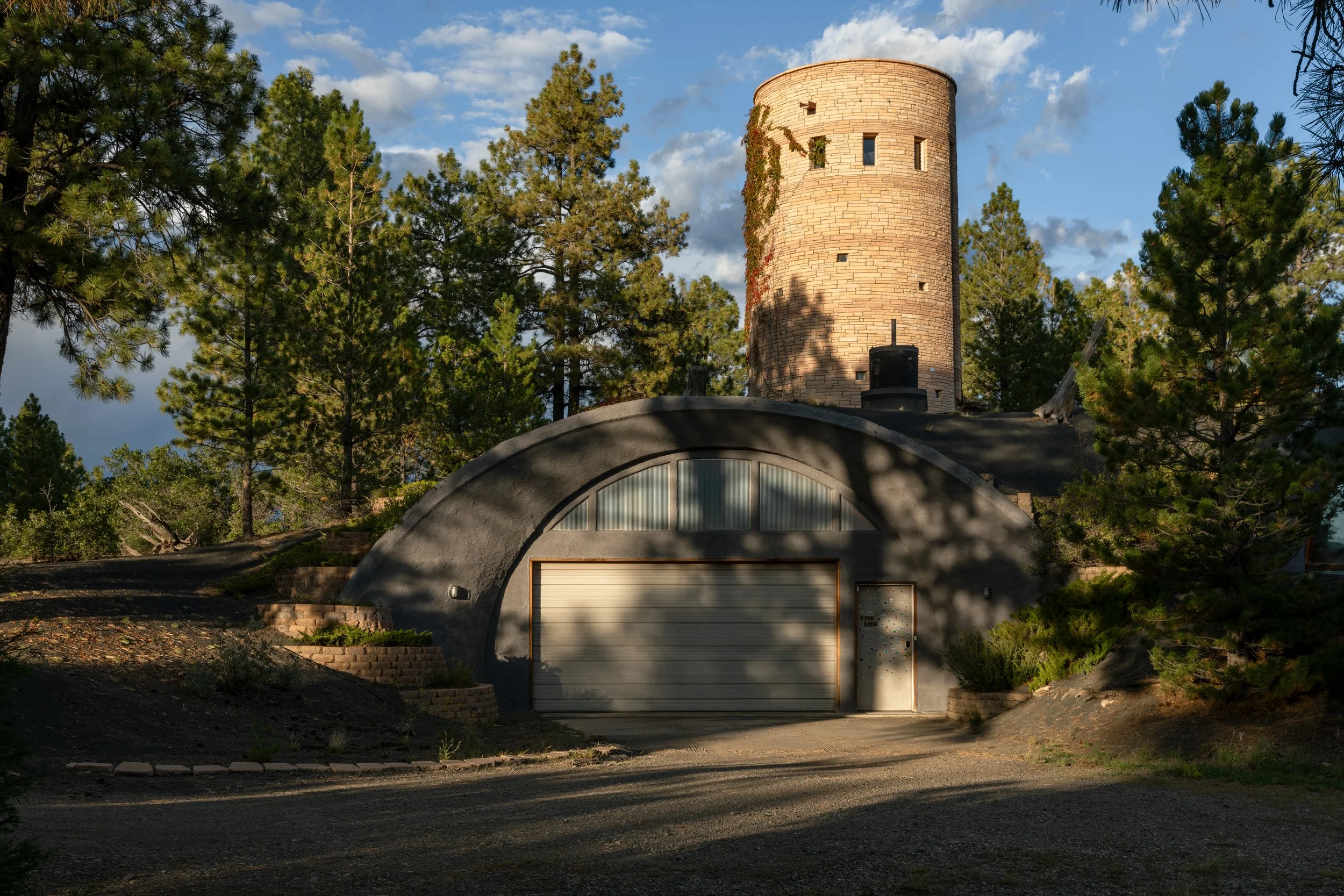 A house built into a hillside with a rounded garage door, surrounded by trees, with a round tower in the background under a partly cloudy sky.
