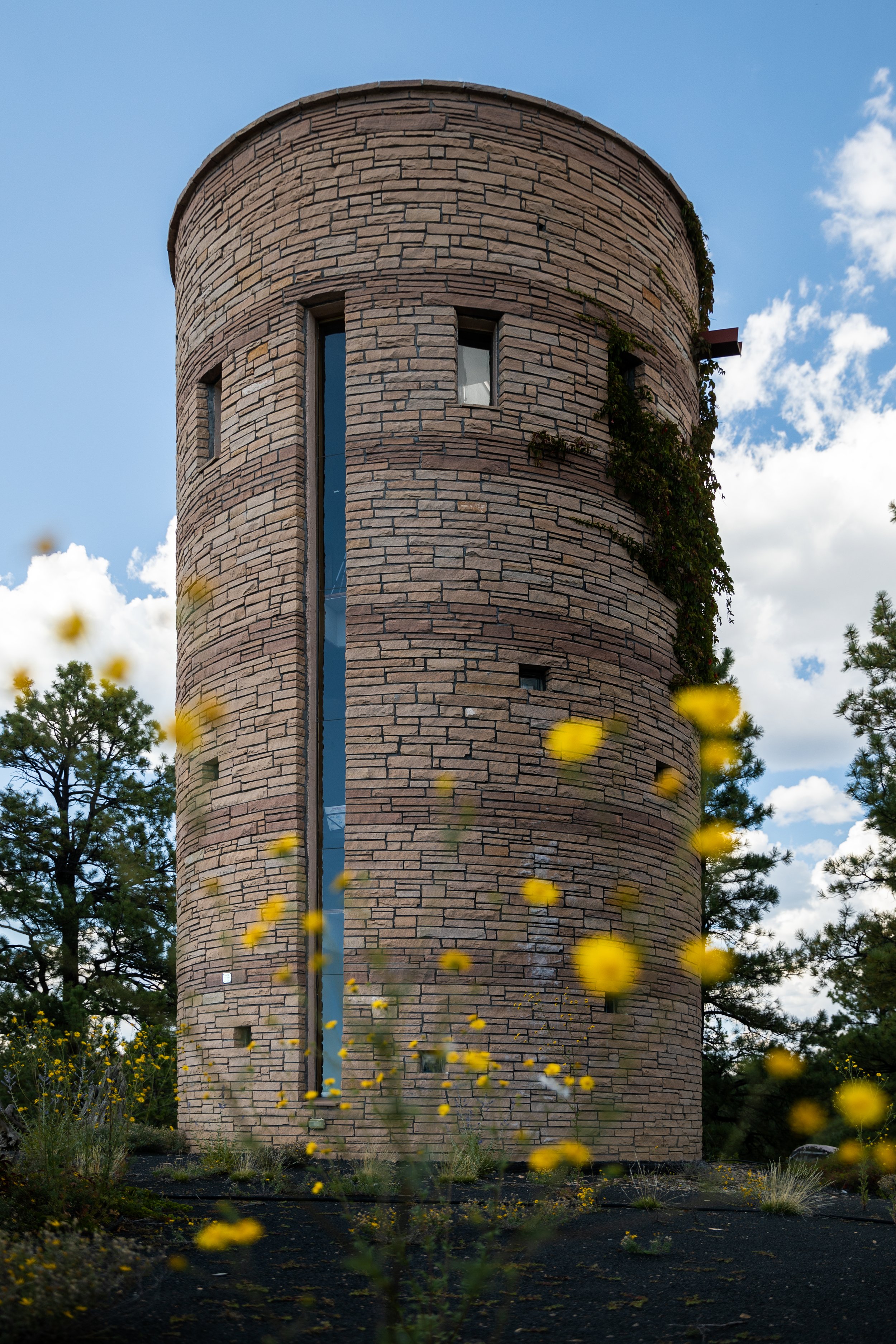 A tall, round brick tower with narrow windows, surrounded by trees and yellow wildflowers, under a partly cloudy blue sky.