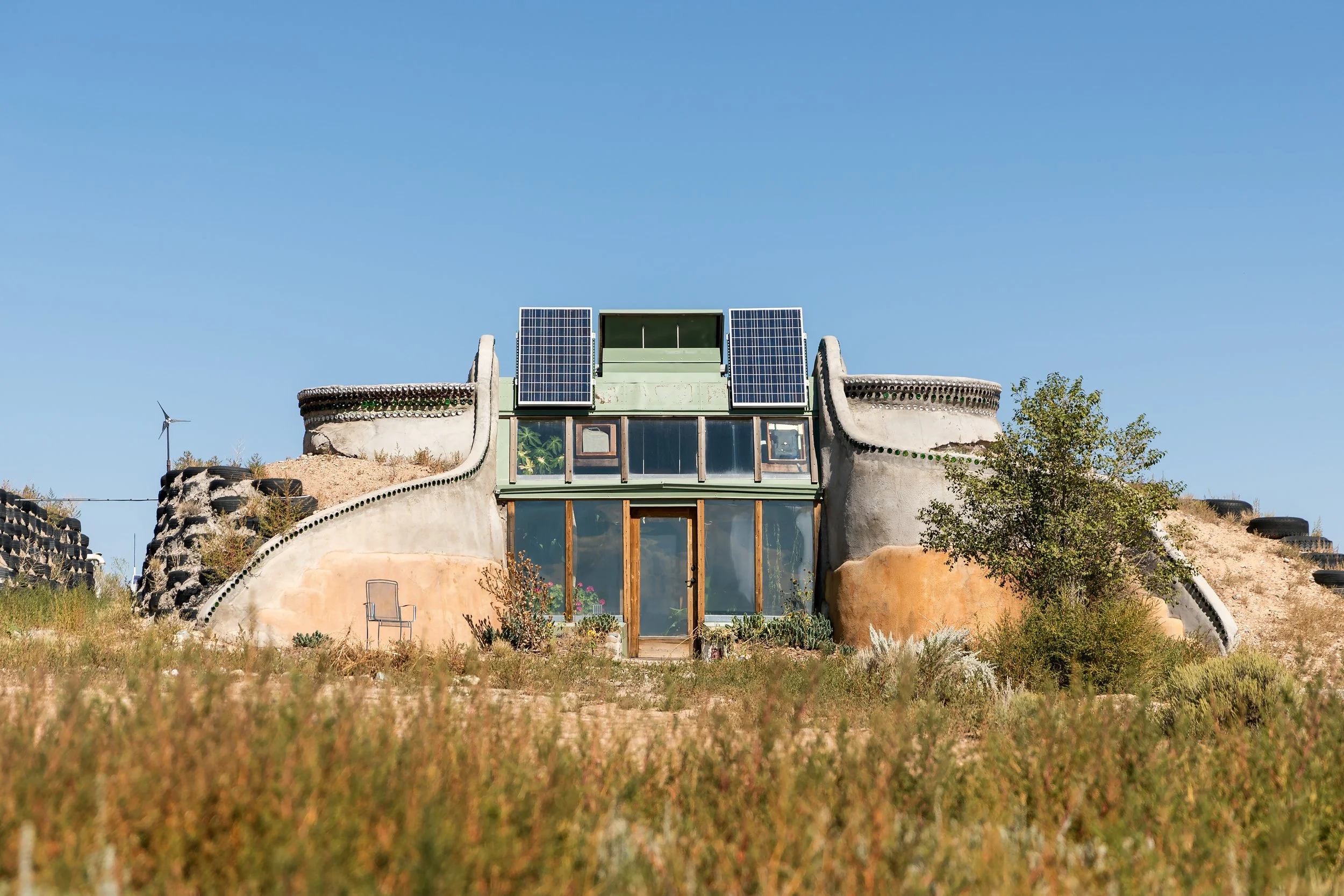 Earthship style eco-friendly house with solar panels on roof, surrounded by natural grass and plants under blue sky.