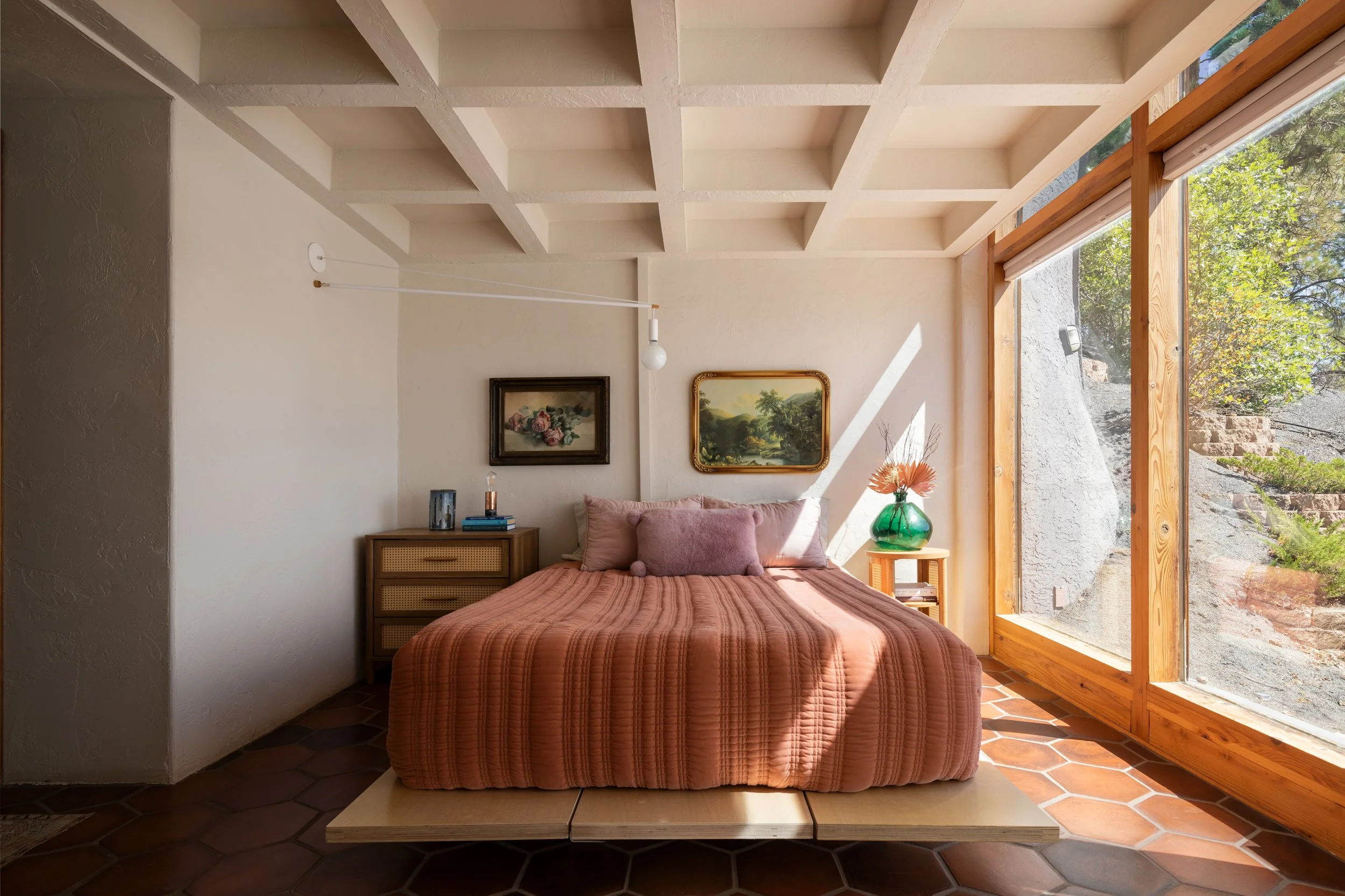 A bedroom with a large window letting in natural light. A bed with pink bedding and pillows is centered against the wall, flanked by two nightstands. Above the bed, two landscape paintings are hung. The room has a unique ceiling with a grid pattern a