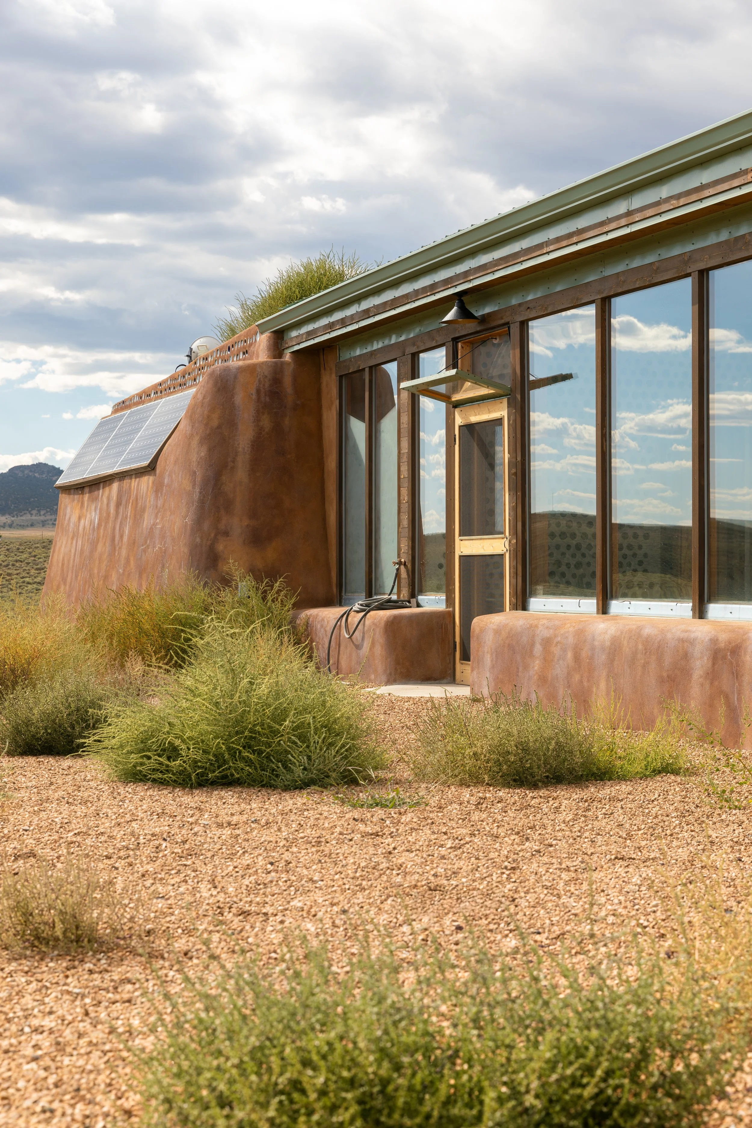 A modern glass and adobe style desert house with solar panels and a gravel yard, set in a semi-arid landscape.