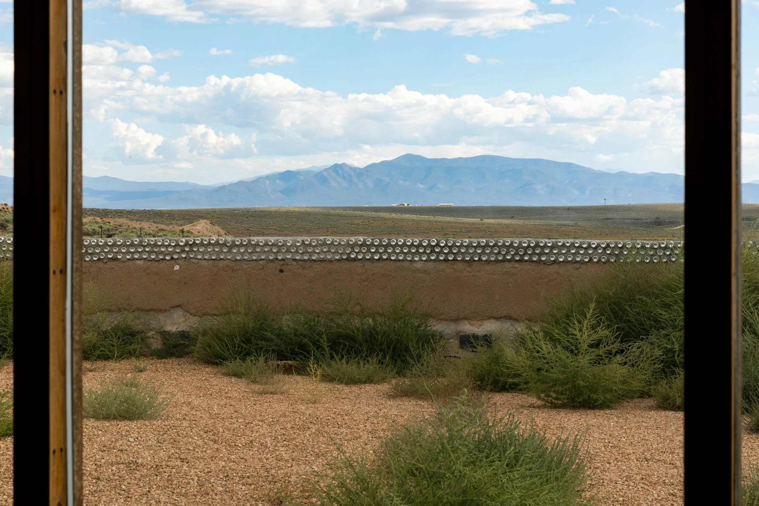 View of a desert landscape with mountains in the distance, seen through a window frame, with a wall made of adobe and metal cans in the foreground.