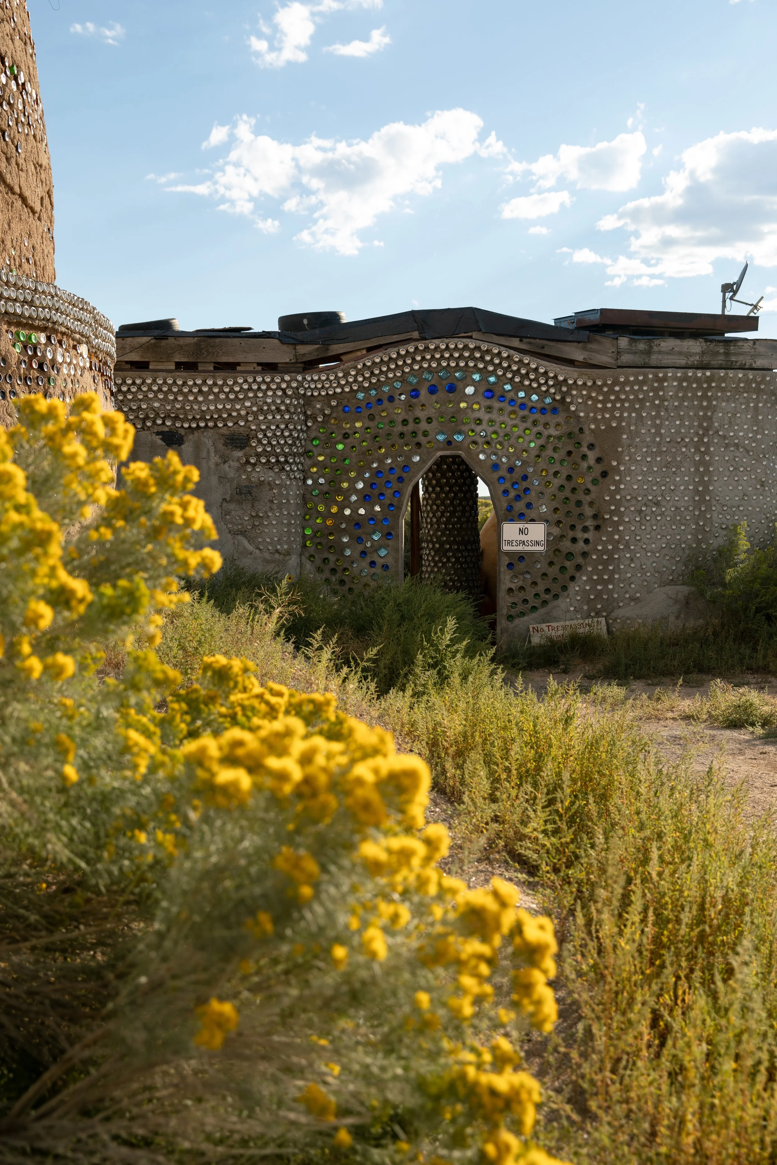 An eclectic building made of concrete and decorated with colorful bottles arranged in patterns, set against a bright sky with scattered clouds. Yellow wildflowers and desert plants grow in the foreground, with a 'No Trespassing' sign on the building.