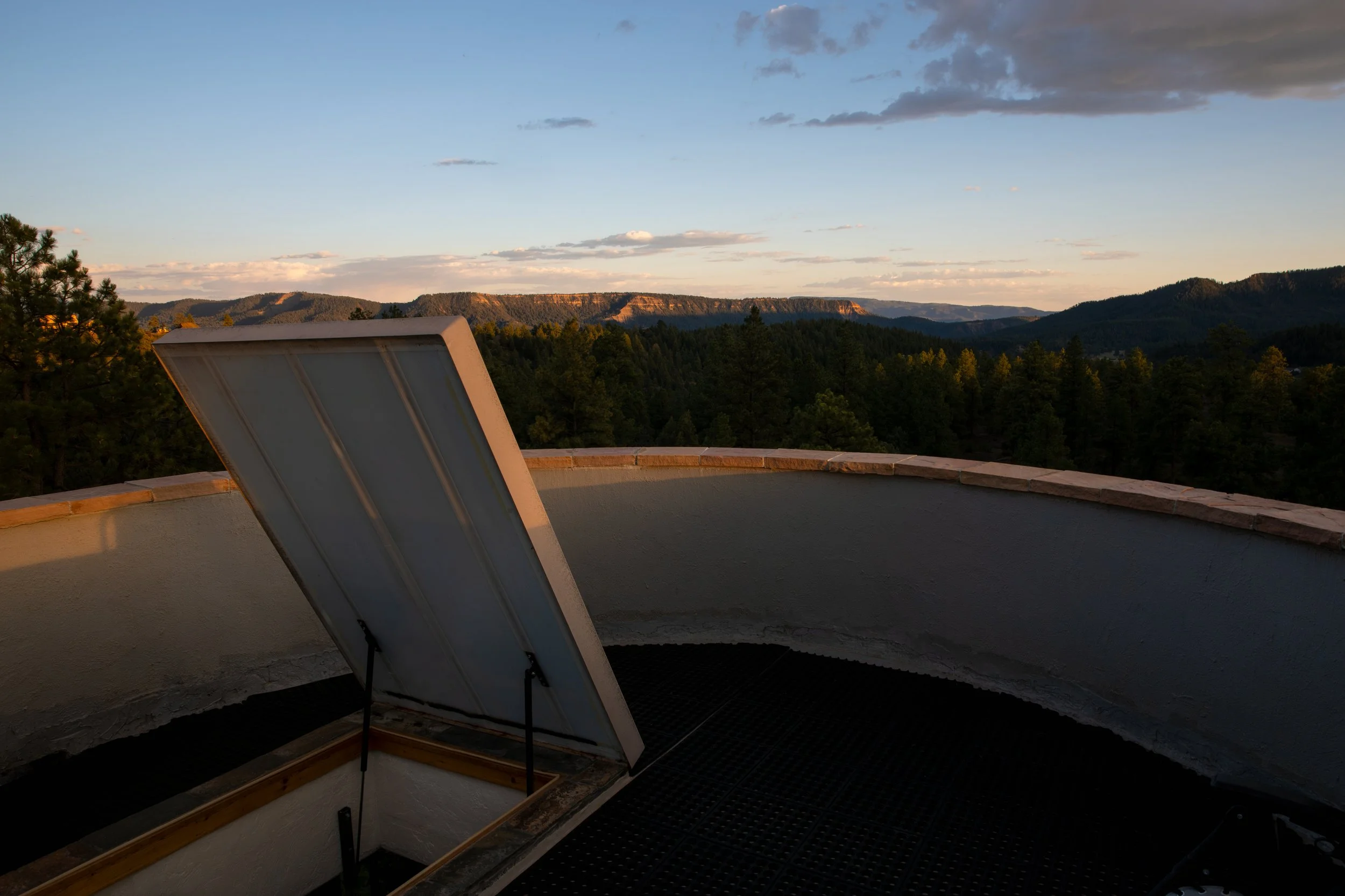 A rooftop telescope hatch open, overlooking a scenic landscape of hills and forests under a partly cloudy sky during sunset.