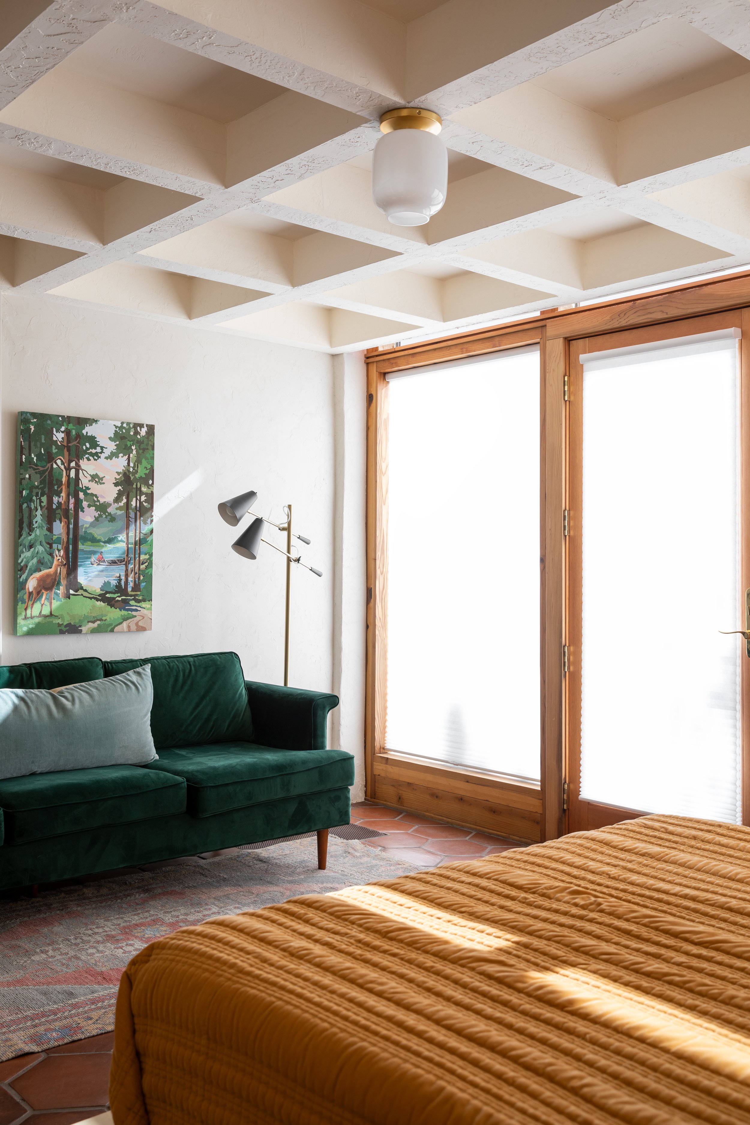 A cozy bedroom with a green velvet couch, a wooden door with window blinds, a piece of artwork of a forest scene, and a bed with orange bedding. Sunlight filters through the windows.