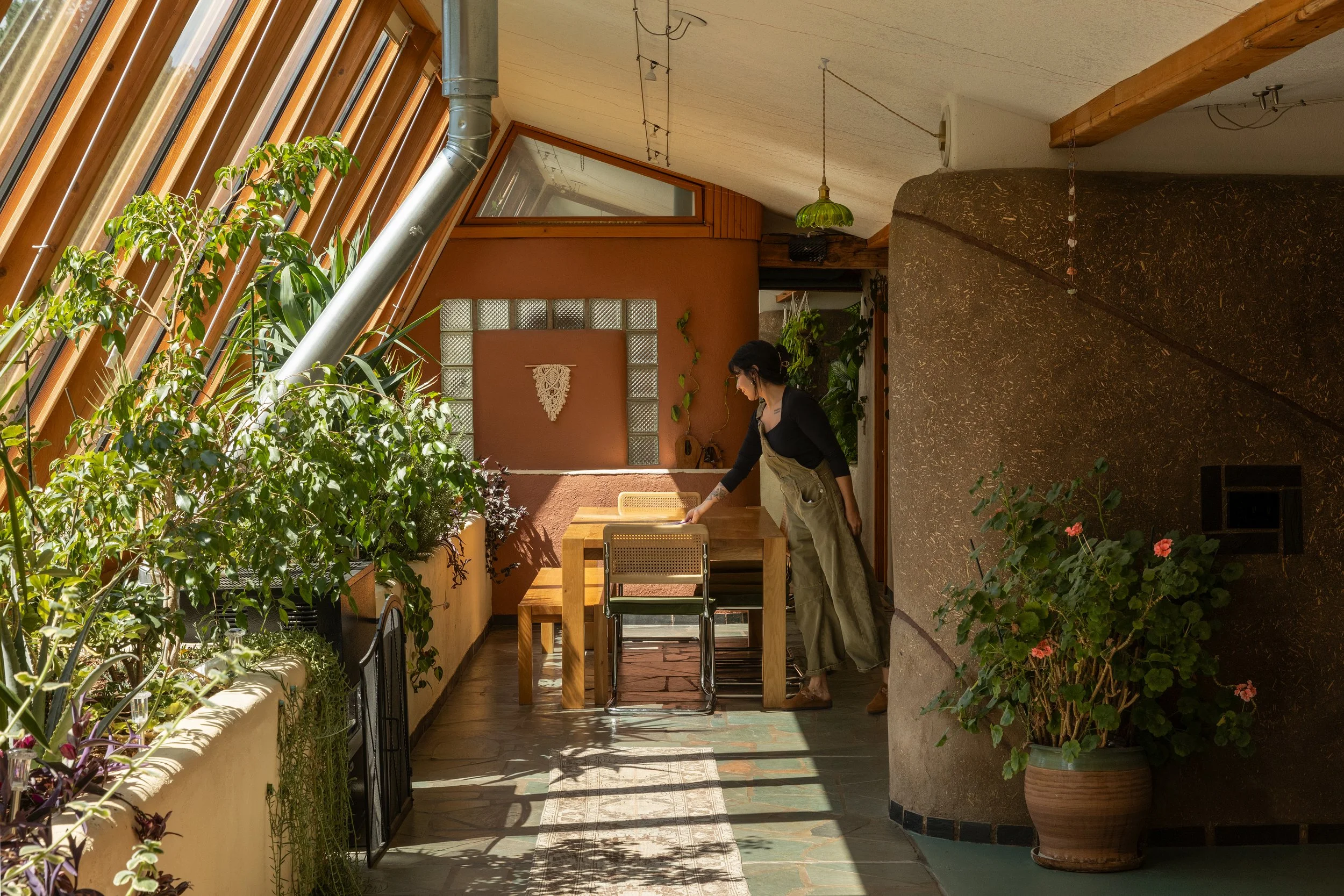 Woman in black shirt and beige apron arranging a wooden dining table in a sunlit indoor garden area with potted plants and colorful decor.