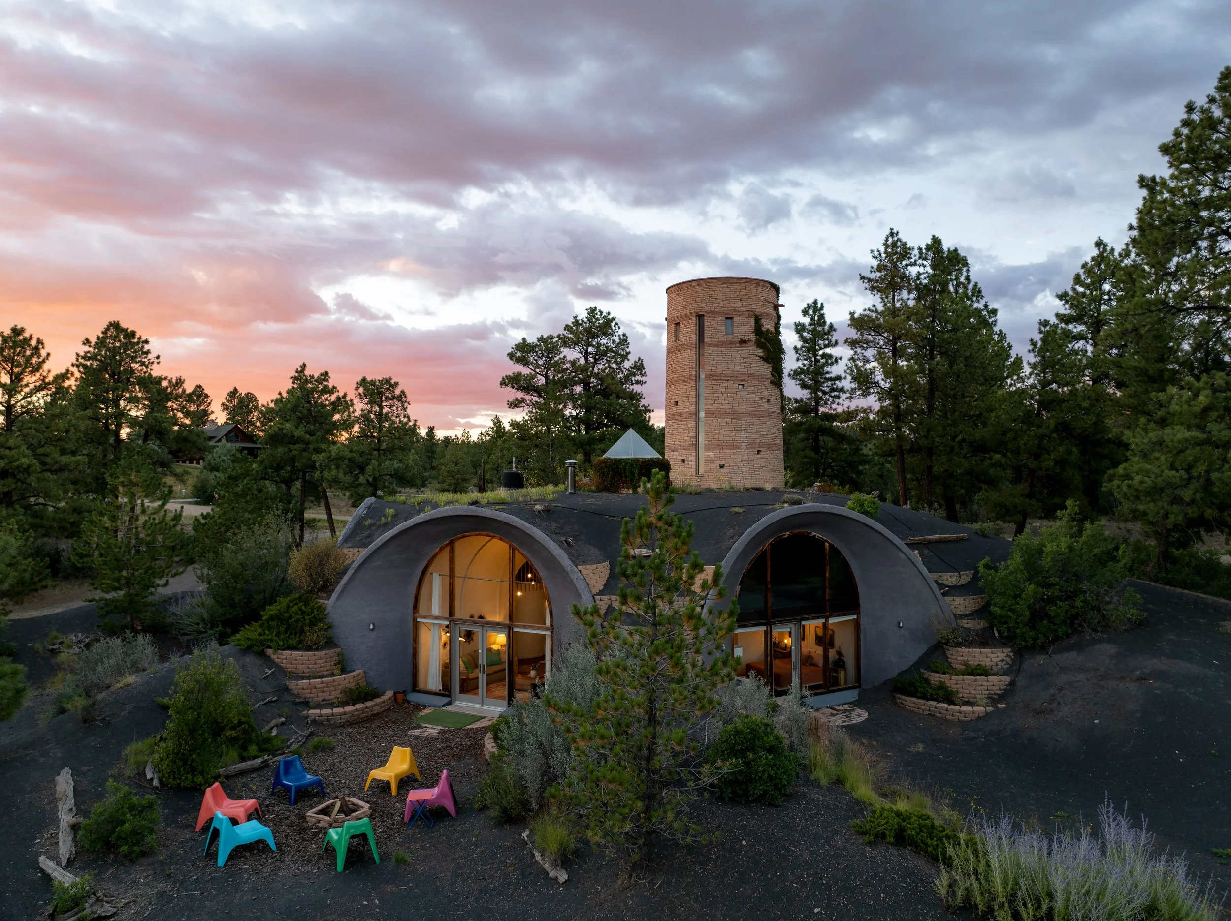 A house with a unique design features a black, curved roof and large, arched windows, situated in a forested area during sunset with a sky filled with clouds. The house includes a small, colorful outdoor seating area with plastic chairs and surrounde