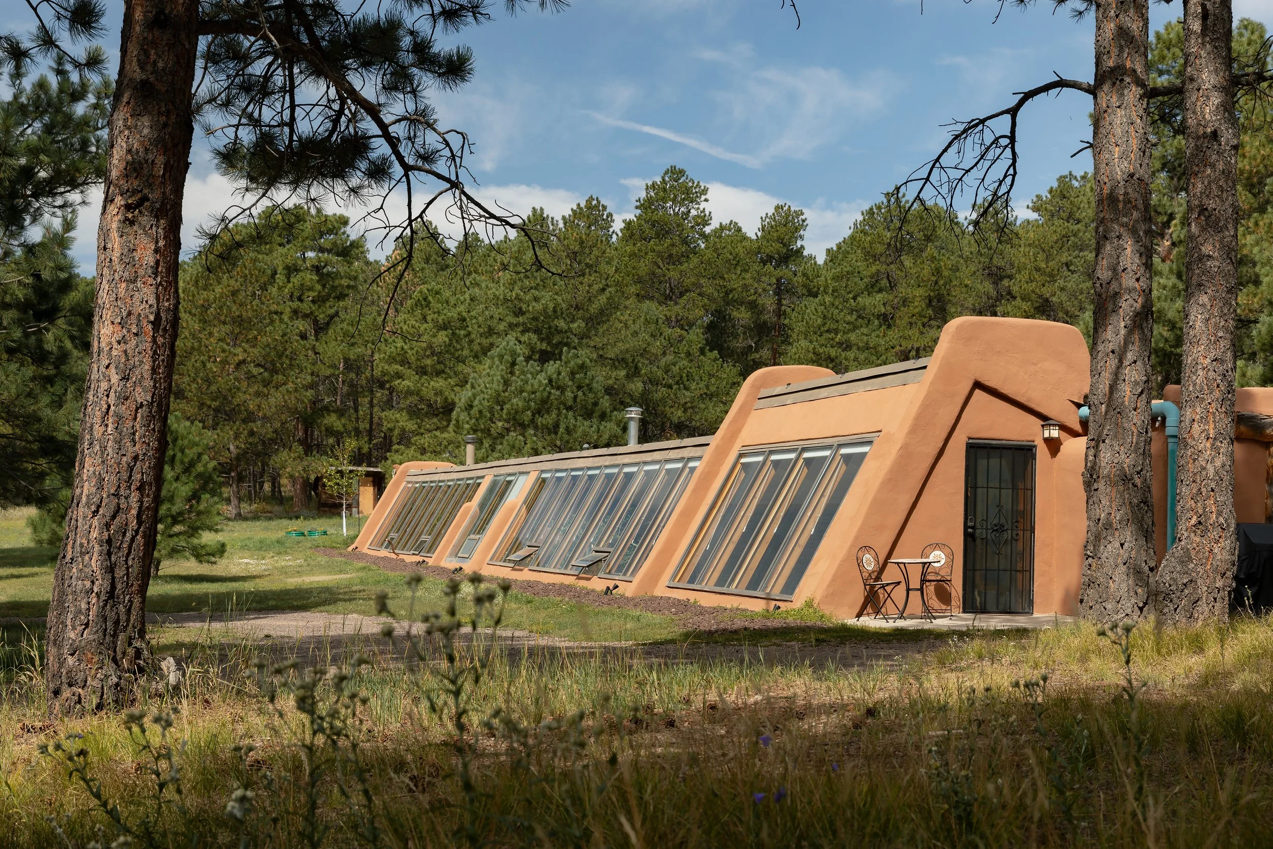 A building with a southwestern style featuring multiple inclined glass skylights, a rounded edge, and a metal security door, surrounded by tall pine trees and a grassy area.
