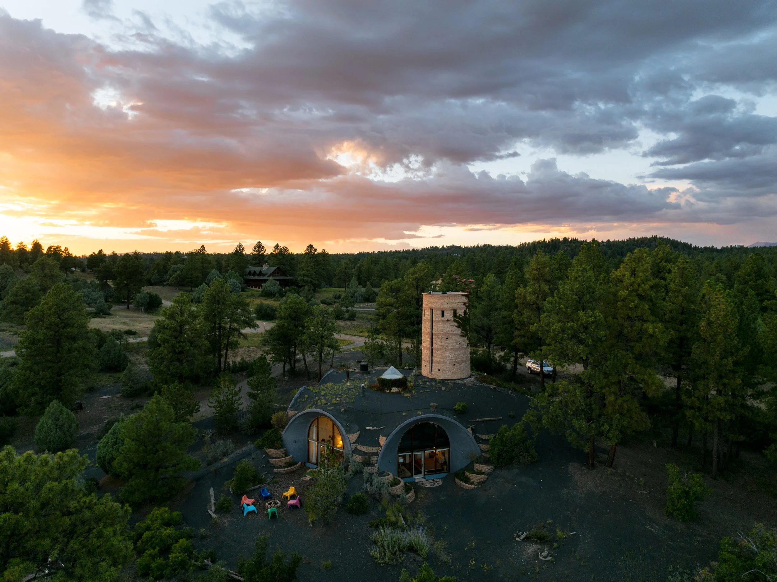 A scenic landscape at sunset featuring a lush forest with a unique house that has curved roofs and large glass doors, surrounded by trees and outdoor chairs.