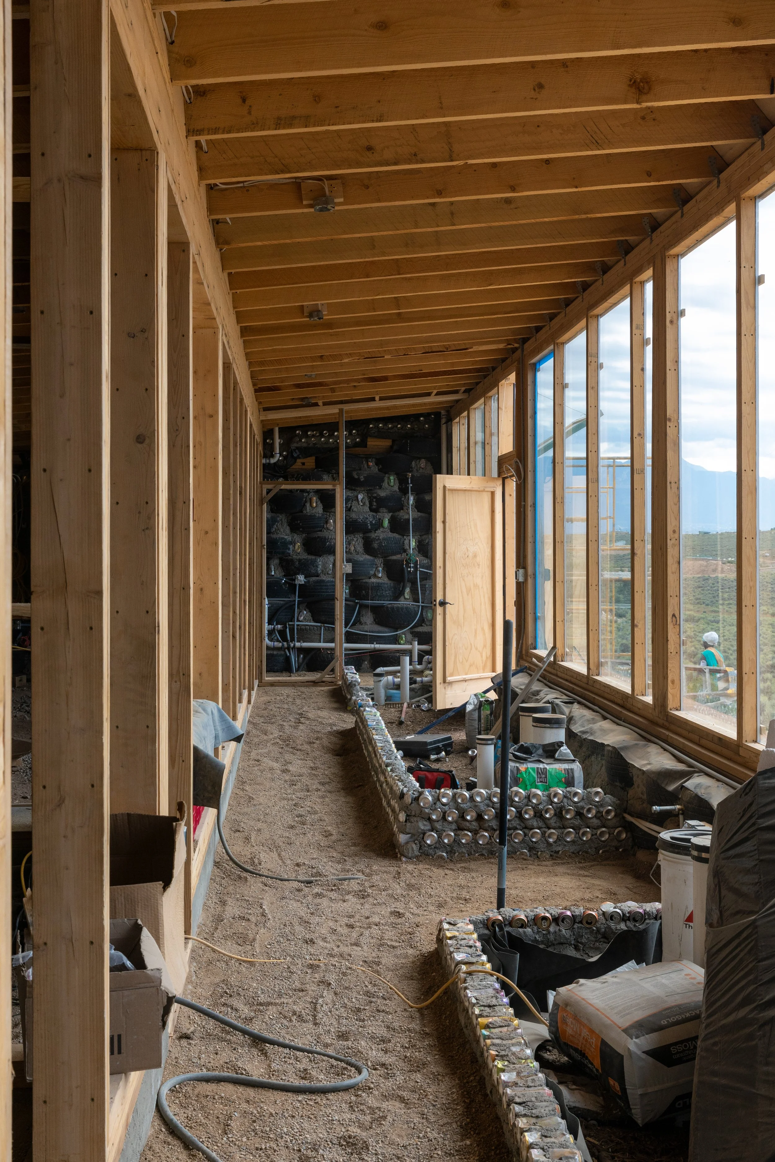 Interior view of a house under construction with exposed wooden framing, a large window on the right, and a sand-covered floor. Construction materials and tools are scattered around.
