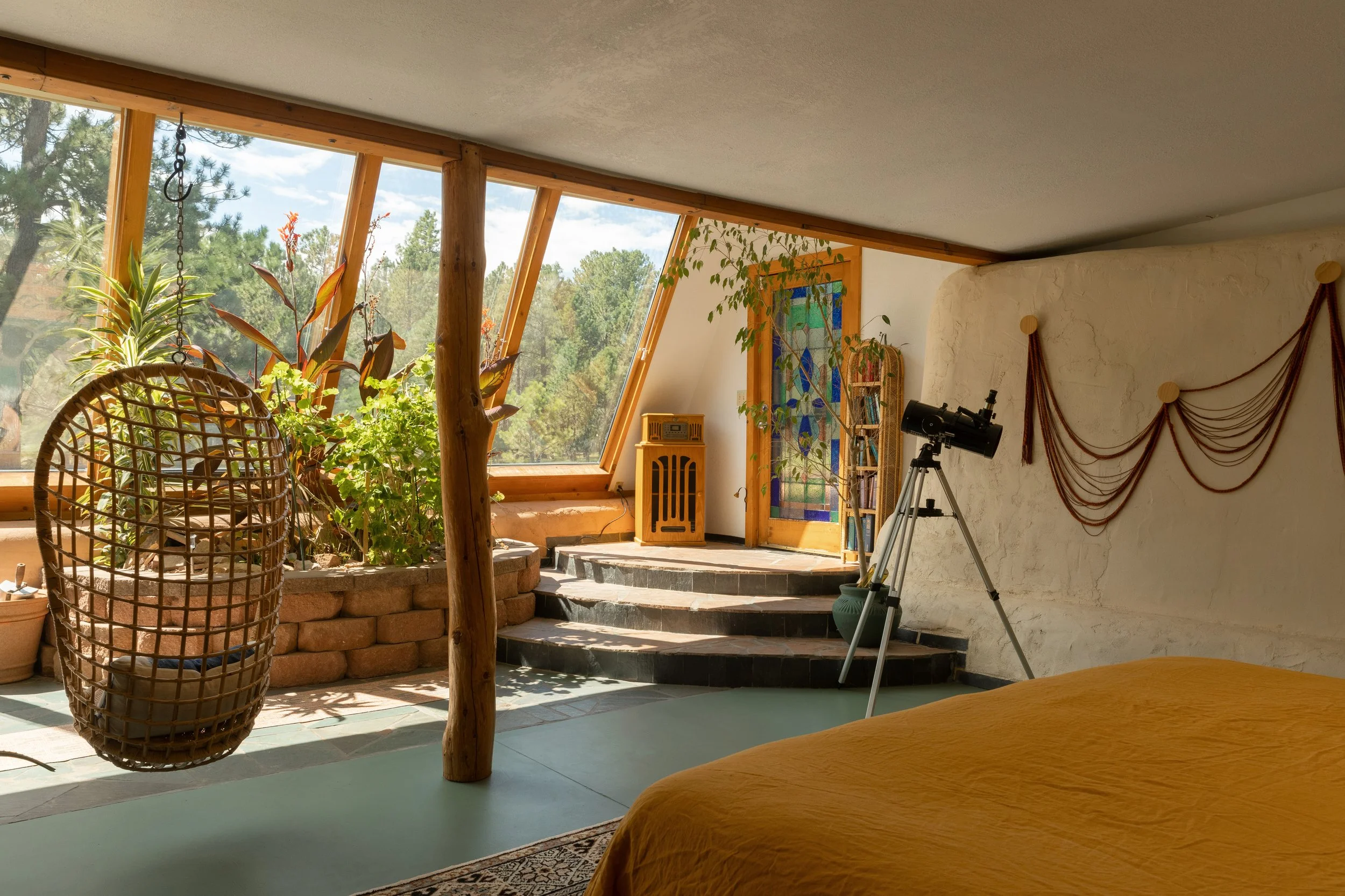 Sunlit room with large angled window, hanging wicker chair, indoor plants, telescope, staircase, and colorful stained glass door.