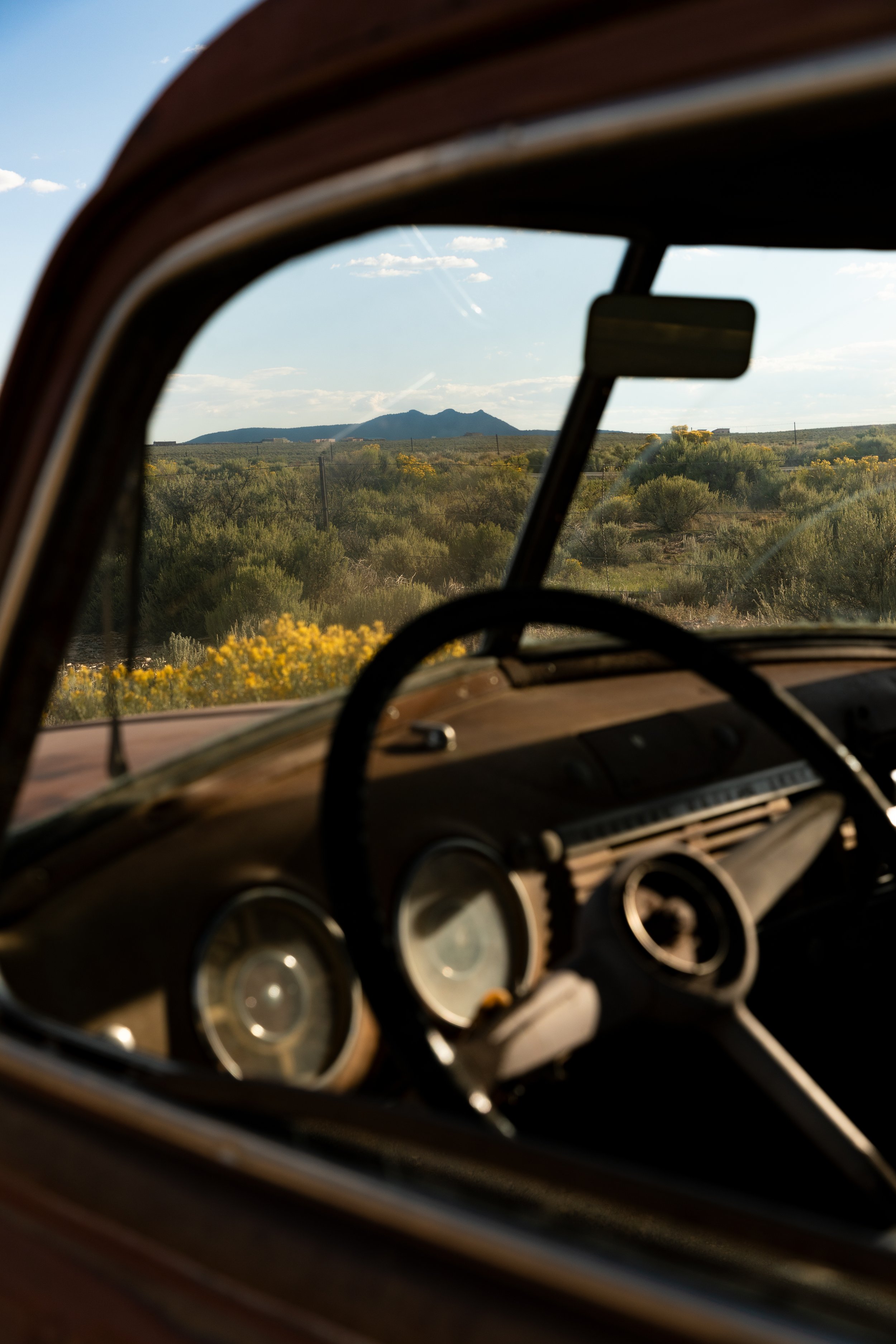 Sunlit interior of a vintage car, showing the dashboard and steering wheel, with a scenic desert landscape and mountains in the distance through the windshield.