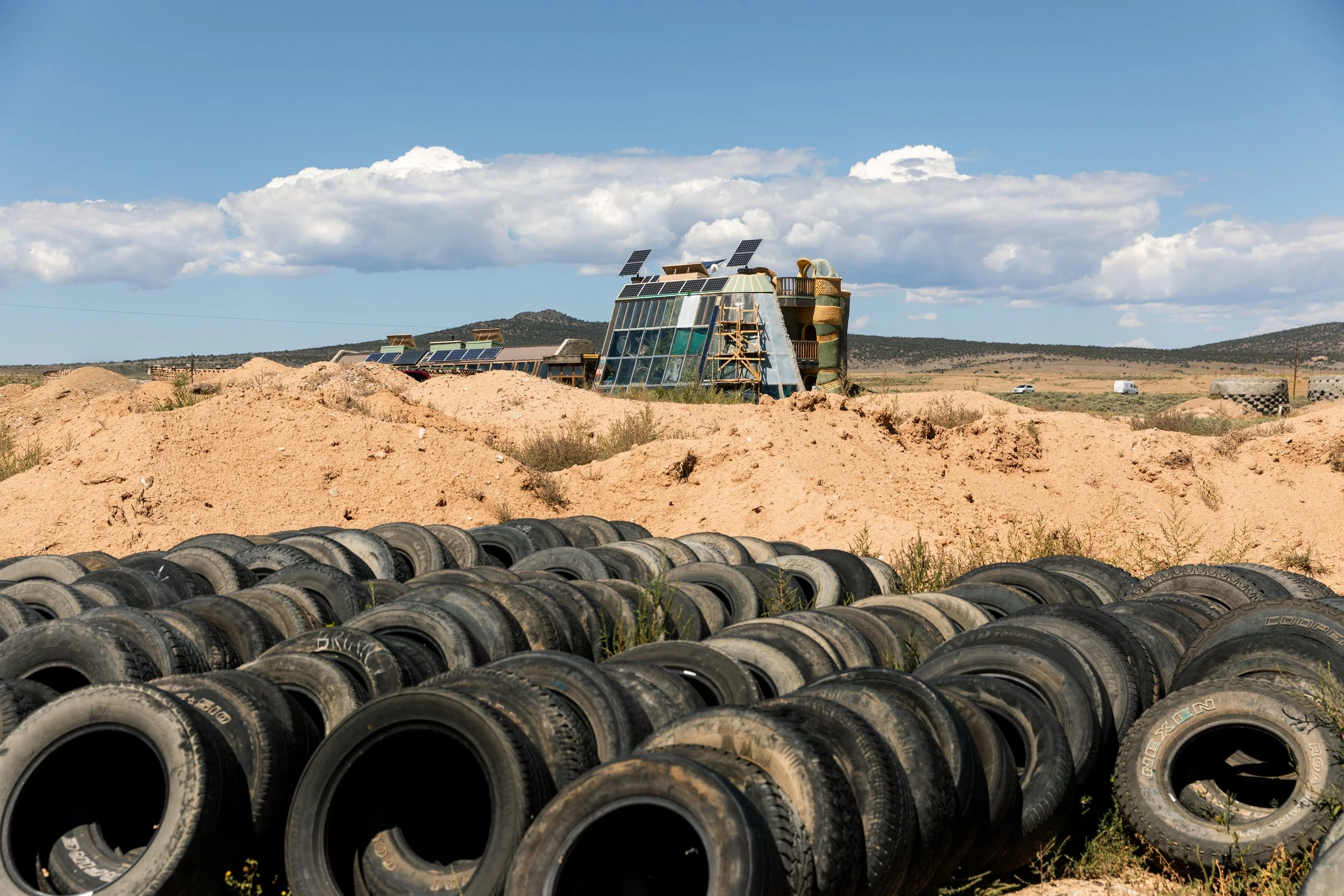 Landscape with piles of used tires in the foreground, a desert-like terrain with a modern building made of glass and wood in the middle ground, and hills with a partly cloudy sky in the background.