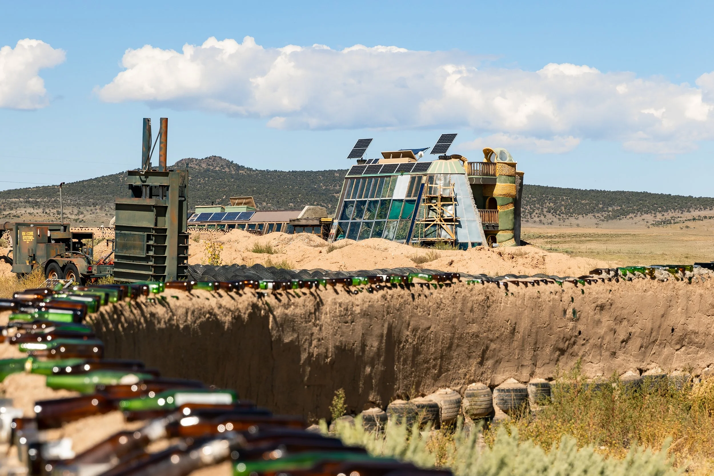 An eco-friendly building with solar panels on the roof, situated in a desert landscape with a mountain in the background, and a row of bottles and a trench in the foreground.