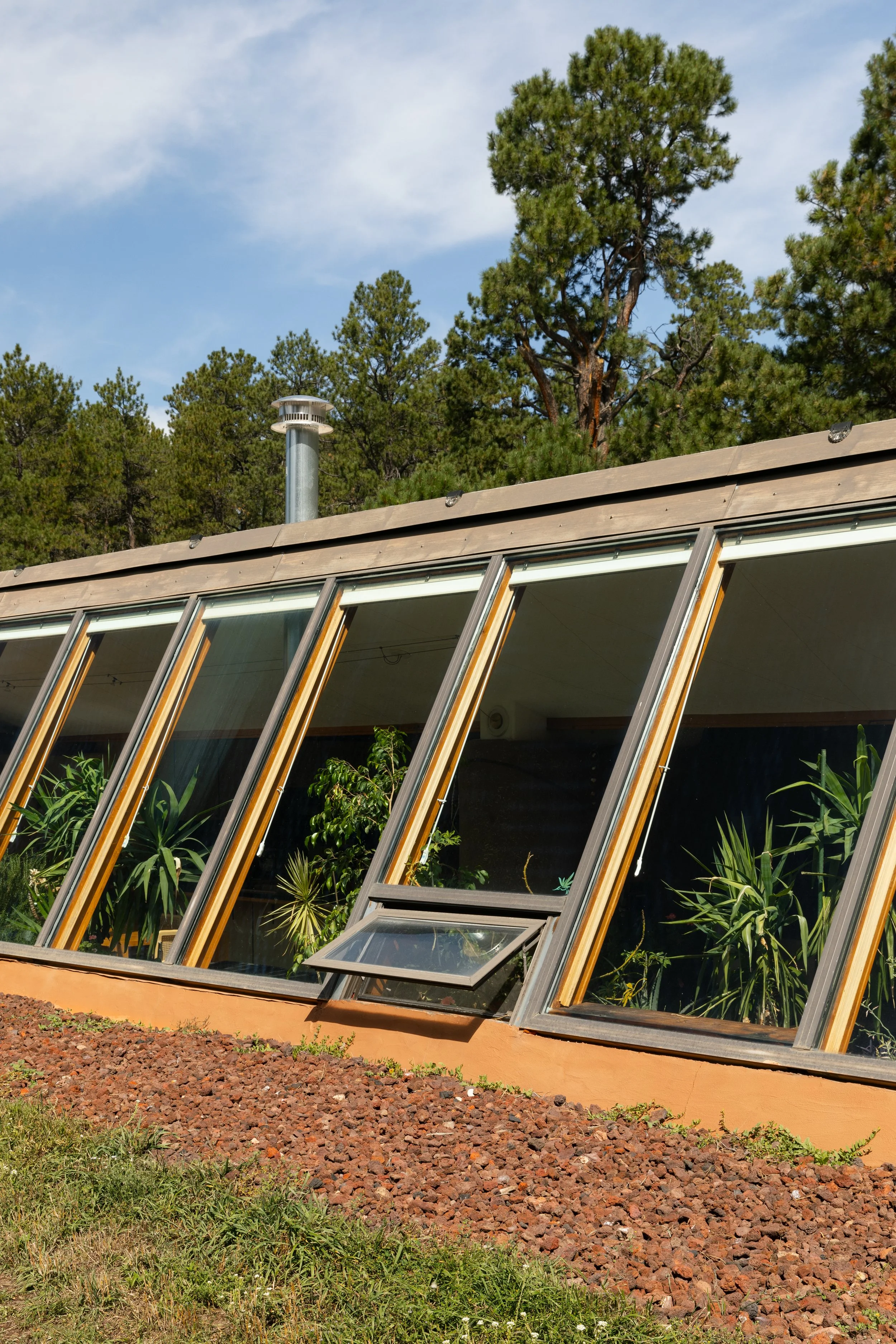 A house with large, angled windows and a rooftop chimney, surrounded by tall green pine trees and a partly cloudy sky.