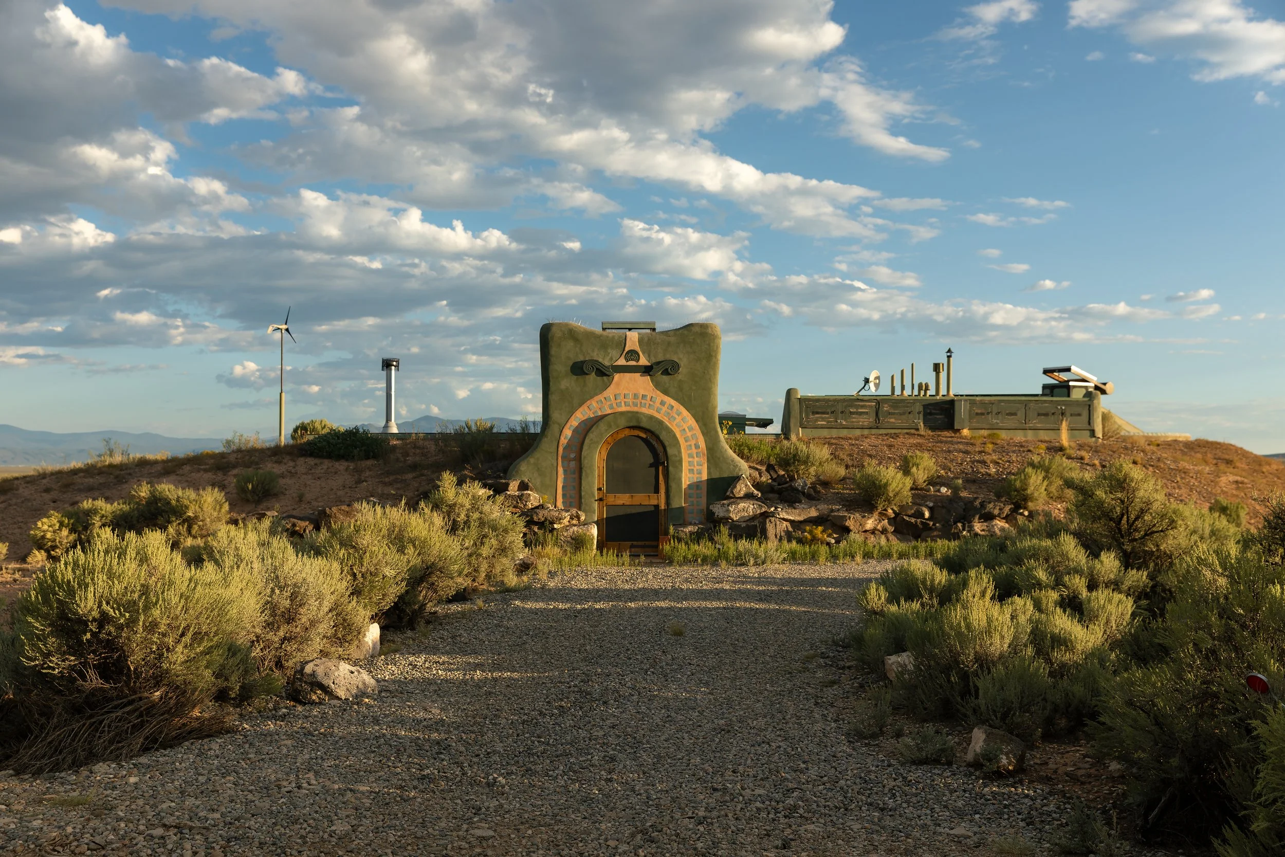 A whimsical, house-like structure with an arched doorway, set in a desert landscape with bushes, a gravel path, an artfully designed chimney, and wind turbines in the background under a partly cloudy sky.
