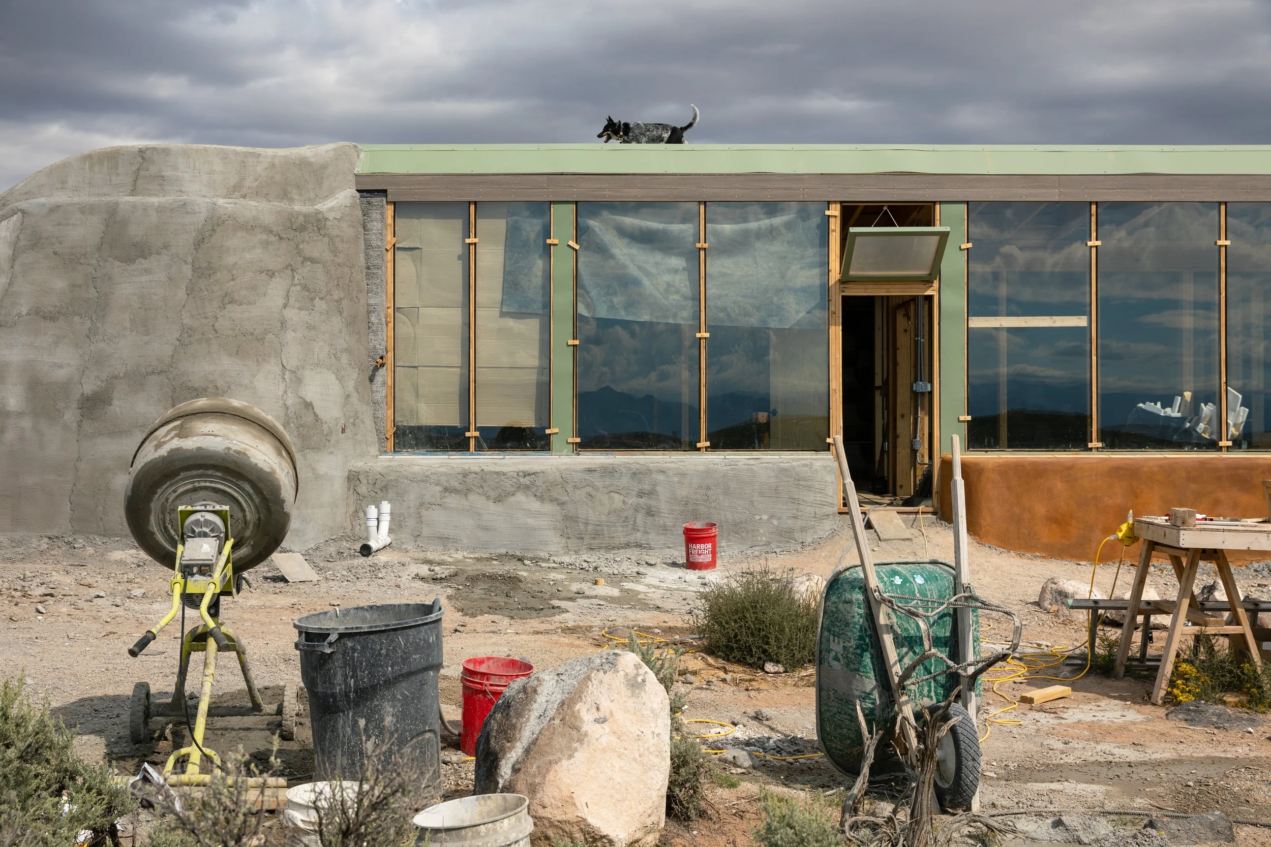 Construction site featuring a modern building with large glass windows, a dog walking on the roof, and construction tools and materials scattered in the foreground under a cloudy sky.