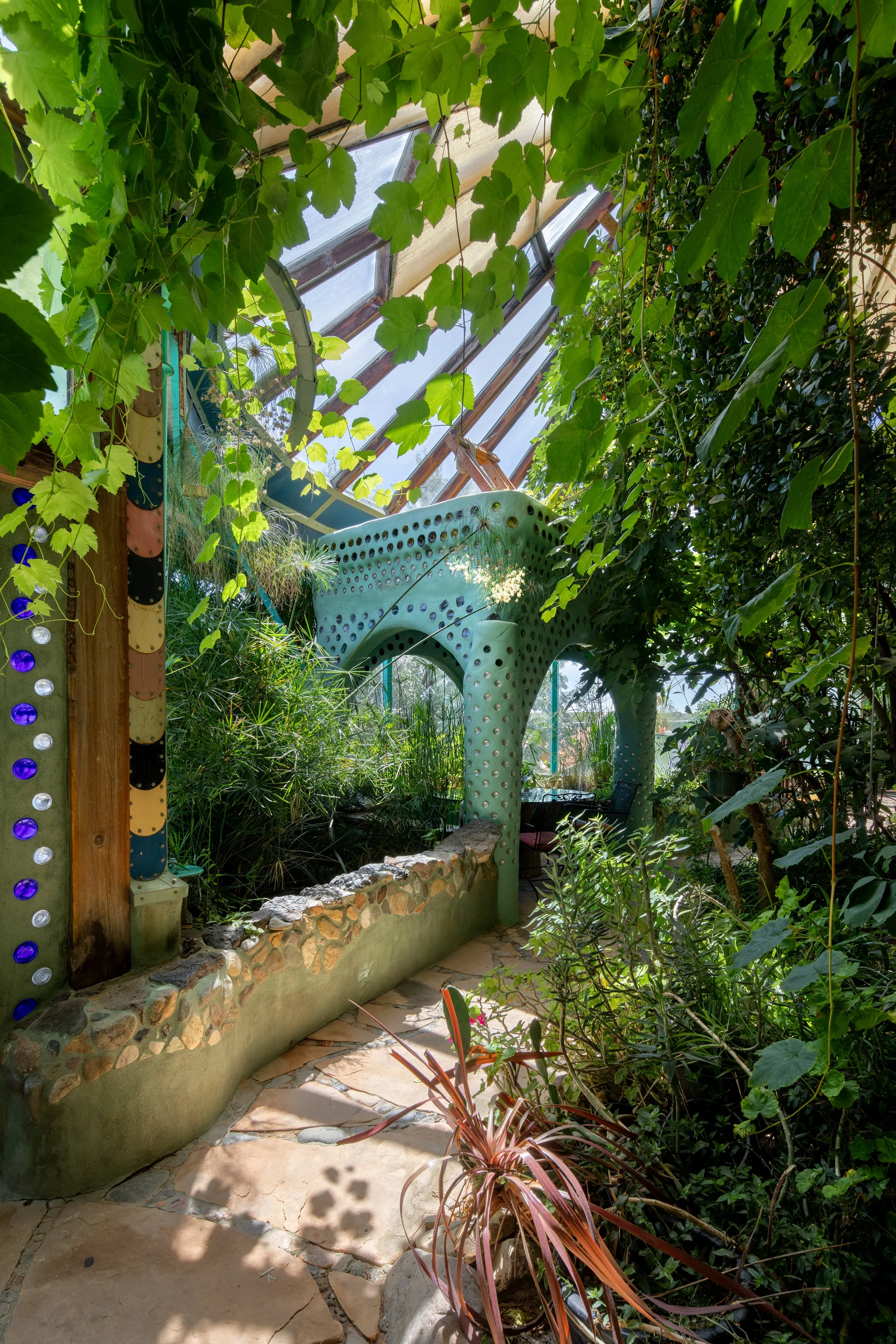 Indoor garden with lush green plants, a greenhouse structure with glass roof, and a decorative blue-green sculpture with perforations.