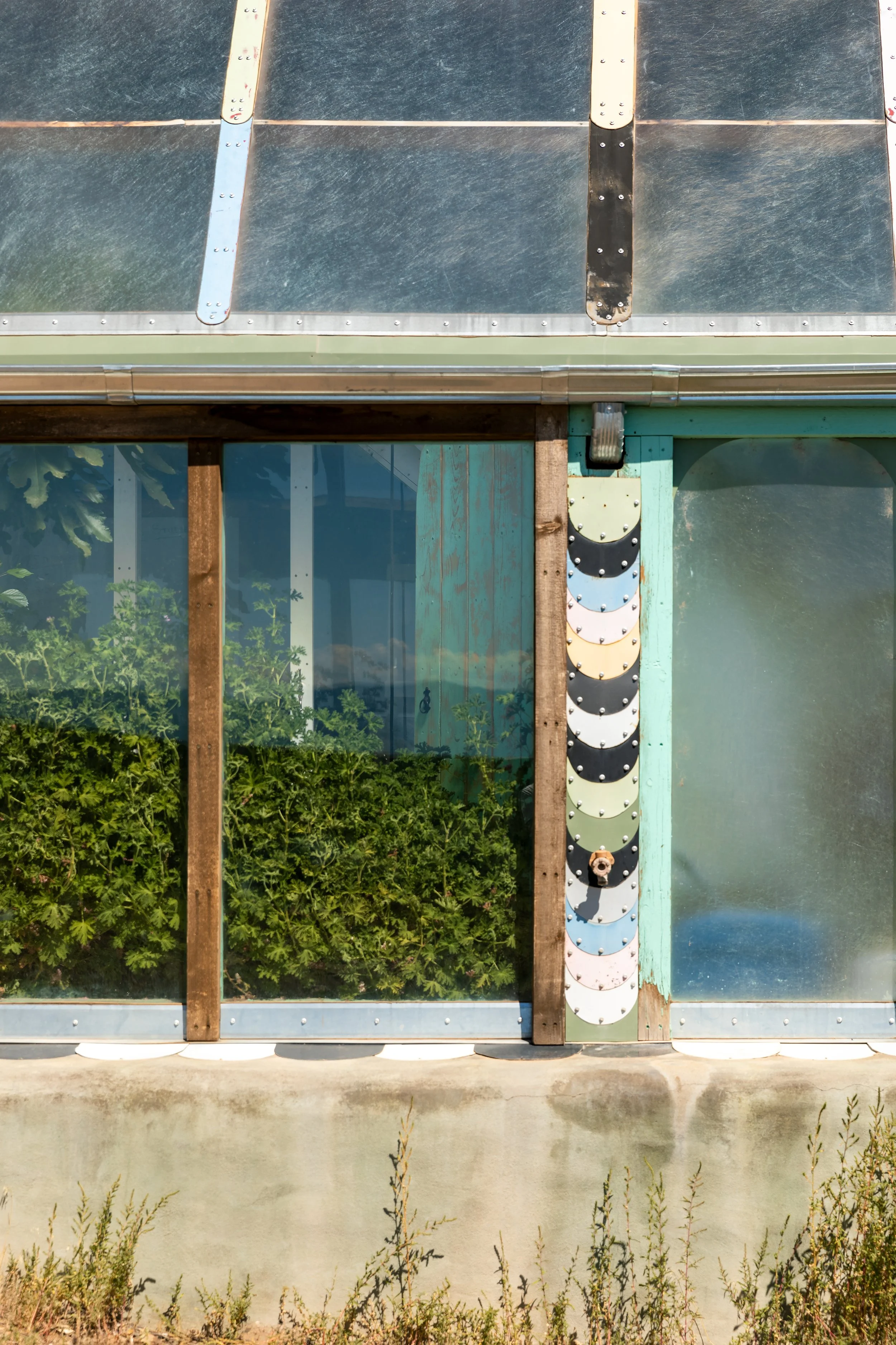 Close-up of the exterior of a greenhouse, showing a glass door, wooden frames, and a vertical row of painted plant labels.