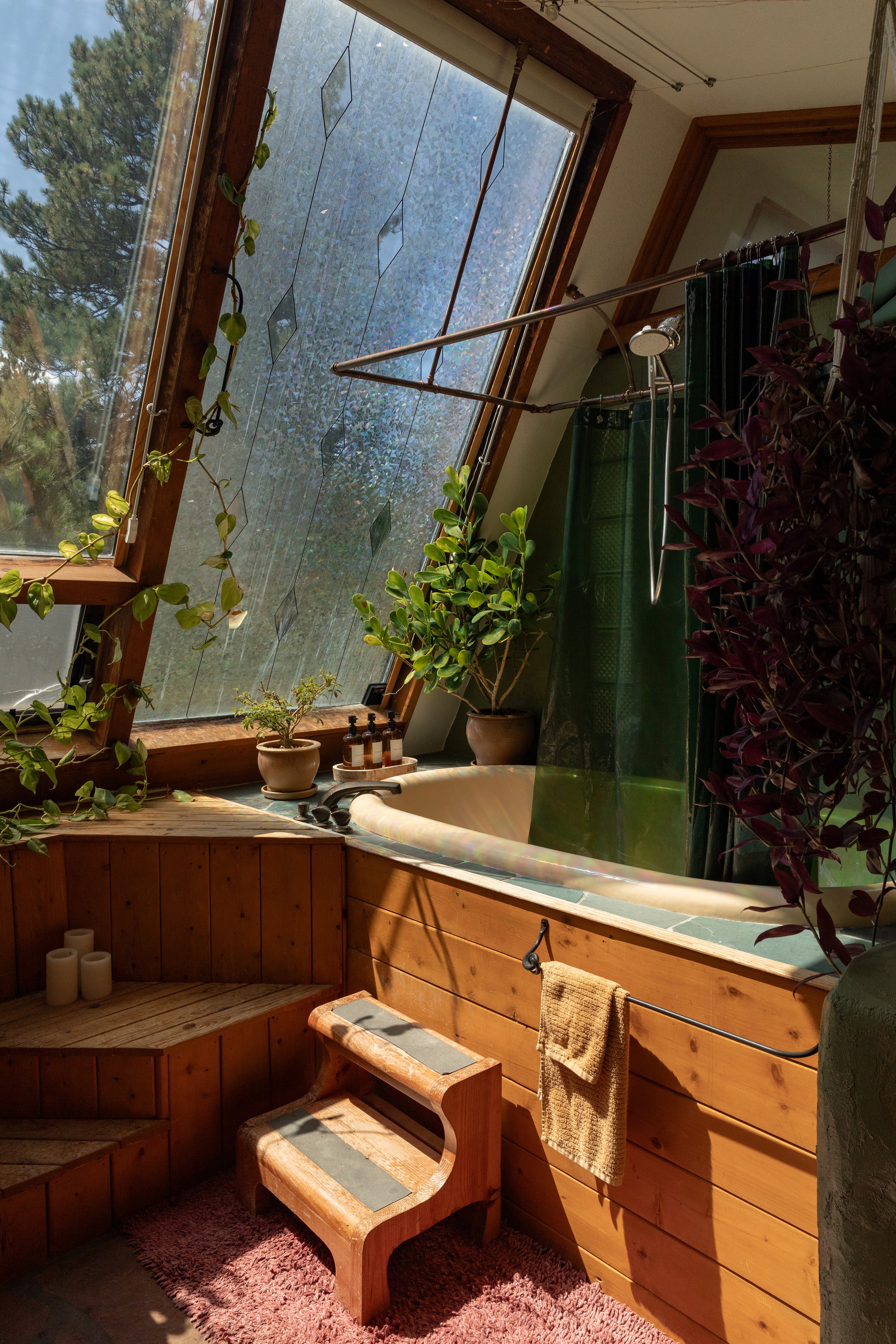 A cozy bathroom with a large sloped window, wooden walls, a built-in bathtub, green curtains, and various potted plants, including a large leafy plant, creating a natural, serene atmosphere.