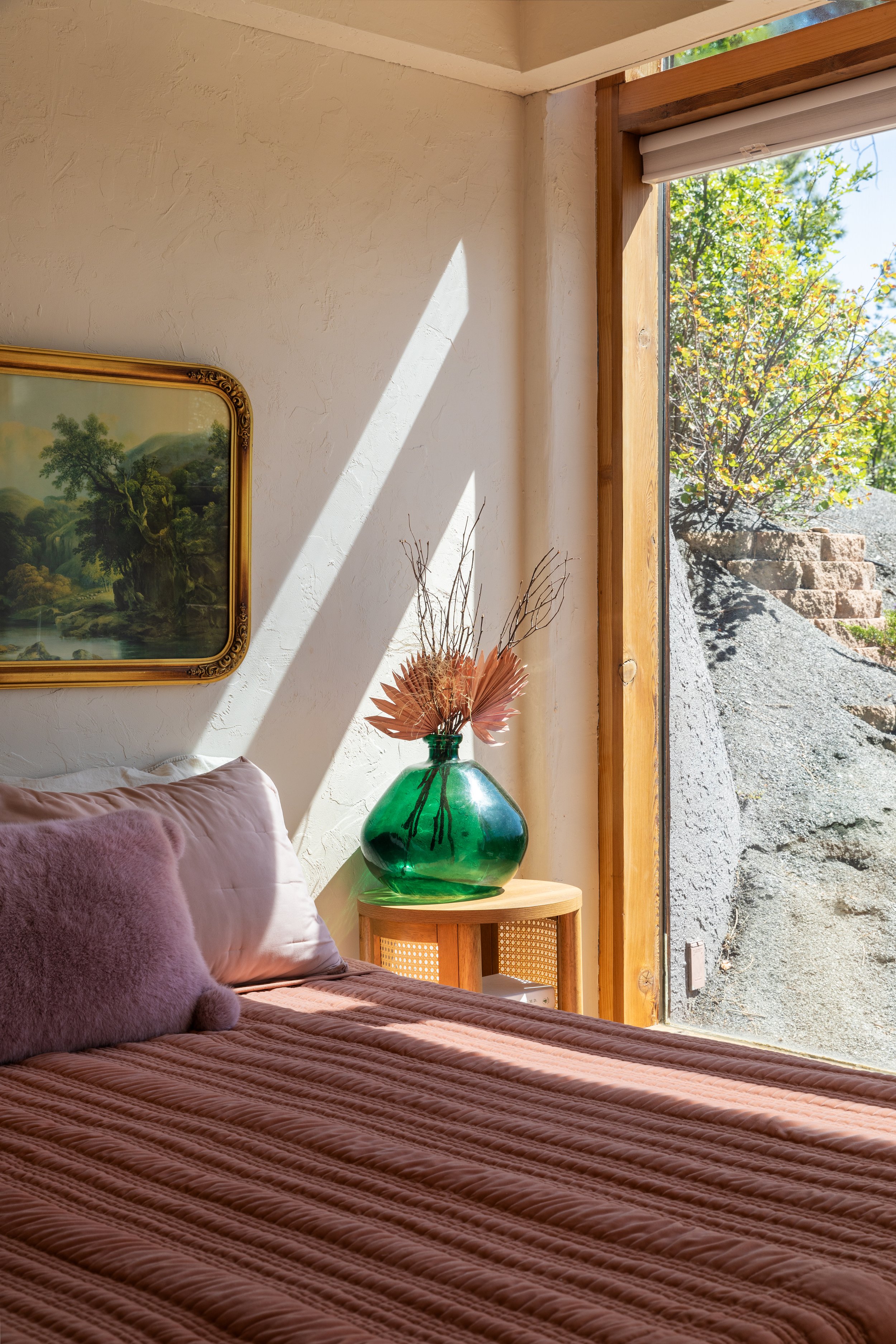 Sunlight streams into a bedroom through large window, illuminating a bed with pinkish-brown textured quilt and two pillows, next to a small wooden side table with a large green glass vase containing dried flowers and branches, and a landscape paintin