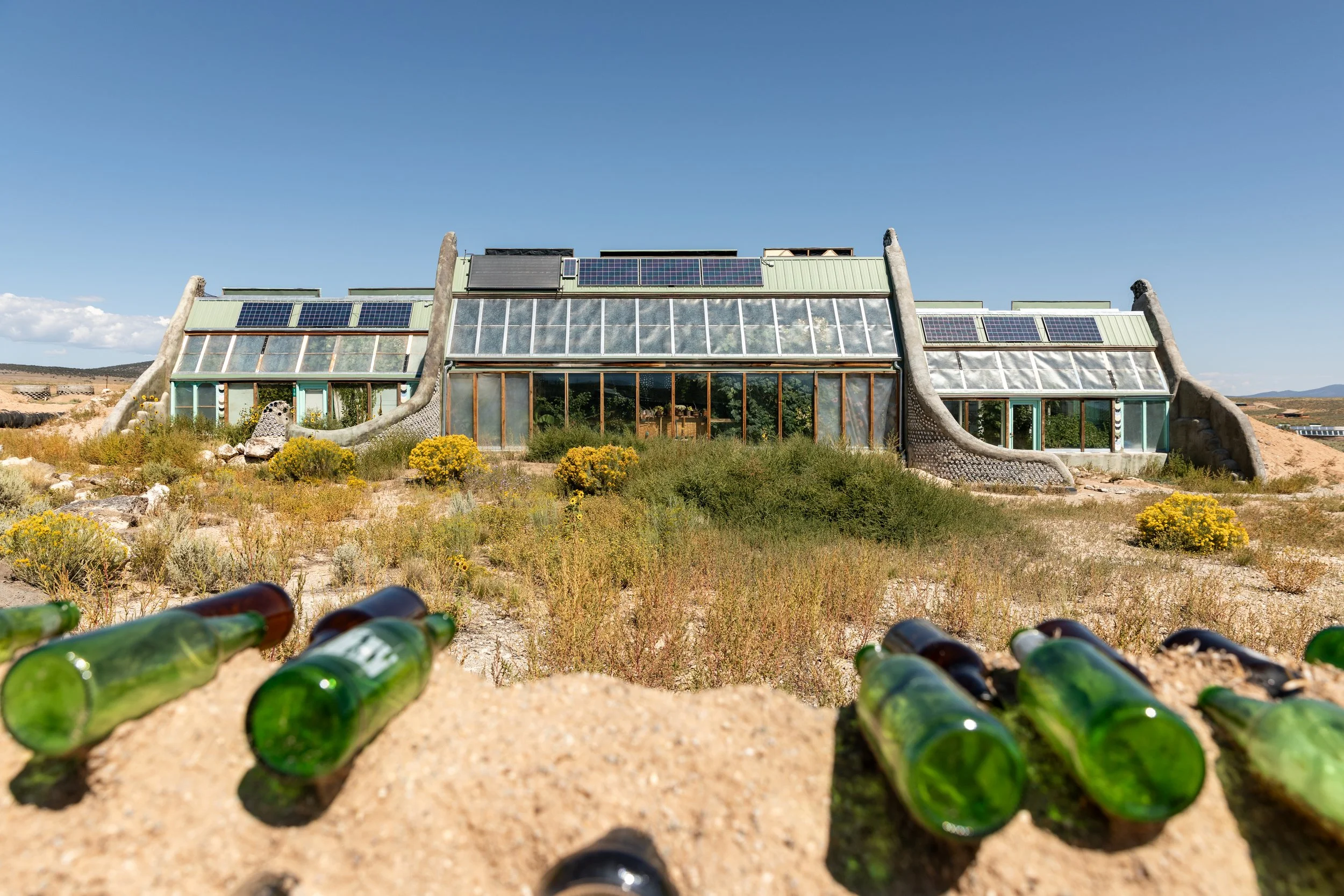 A unique house built in a desert landscape with glass walls and tentacle-like structures on both sides, surrounded by yellow bushes, with green glass bottles on the ground in the foreground.