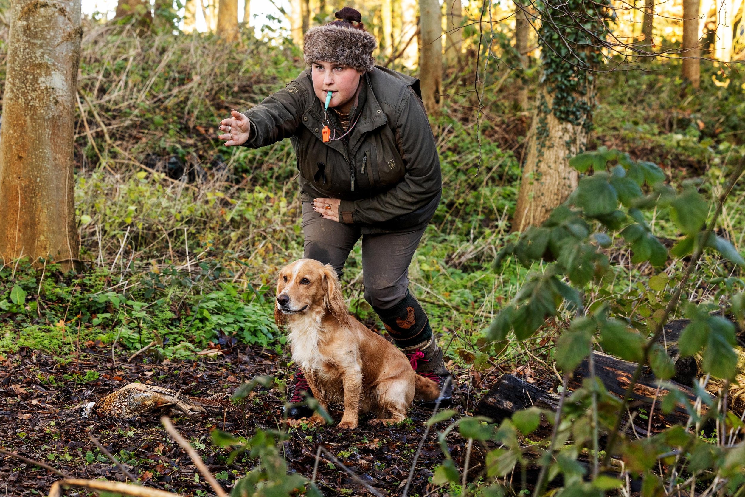A person in outdoor clothing with a whistle around their neck is pointing forward in a wooded area, with a golden retriever sitting beside them on the forest floor.