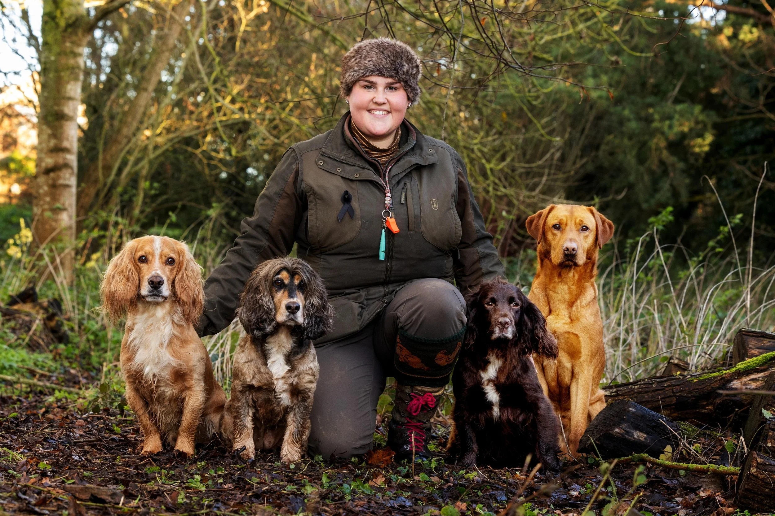 A woman kneeling outdoors in a forest with five dogs, all sitting on the ground. The woman is wearing a brown hat, dark jacket, and pants, smiling at the camera. The dogs are of various breeds and colors, including tan, black, and brown, and are look