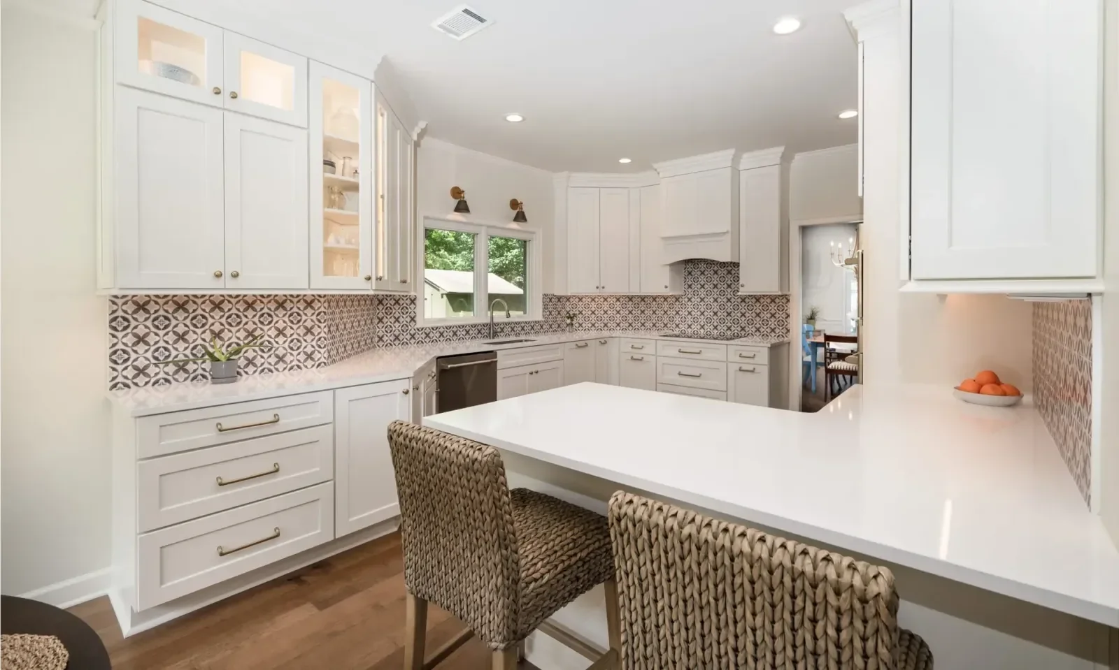 Bright white kitchen with patterned backsplash, white cabinets, and a large island with woven chairs, window with greenery outside.