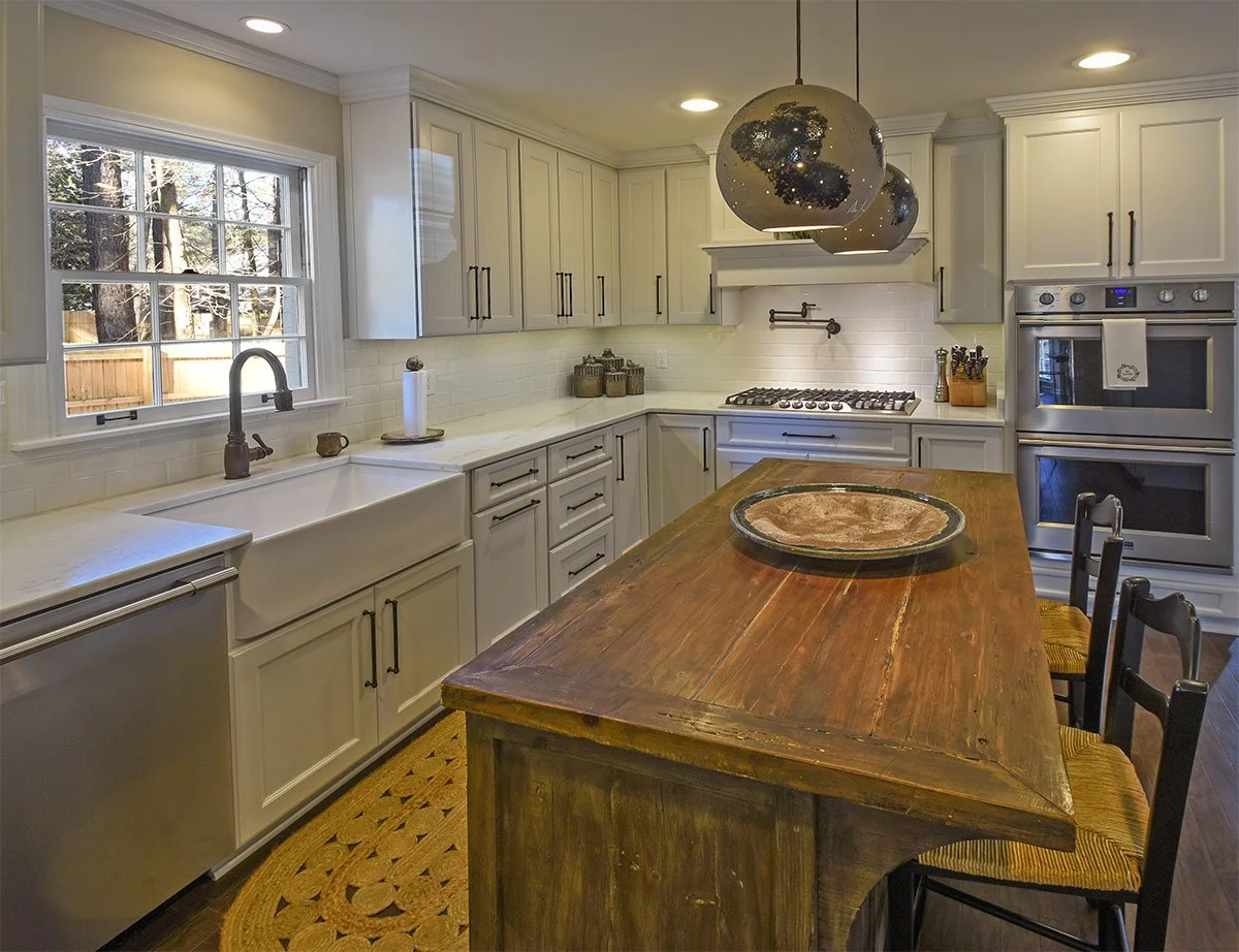 Kitchen with white cabinets, a large window with a view of trees, a farmhouse sink, a wooden island with chairs, and modern appliances.