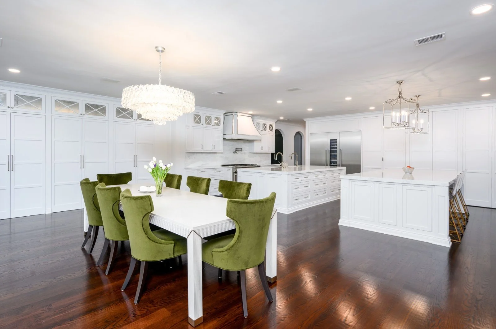 Bright kitchen with white cabinets, marble countertops, and dark hardwood floors. Features a dining table with eight green velvet chairs, a large chandelier, and modern lighting fixtures.