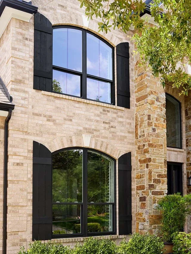 View of a brick house exterior with two large arched windows with black frames and shutters, surrounded by green trees and bushes.