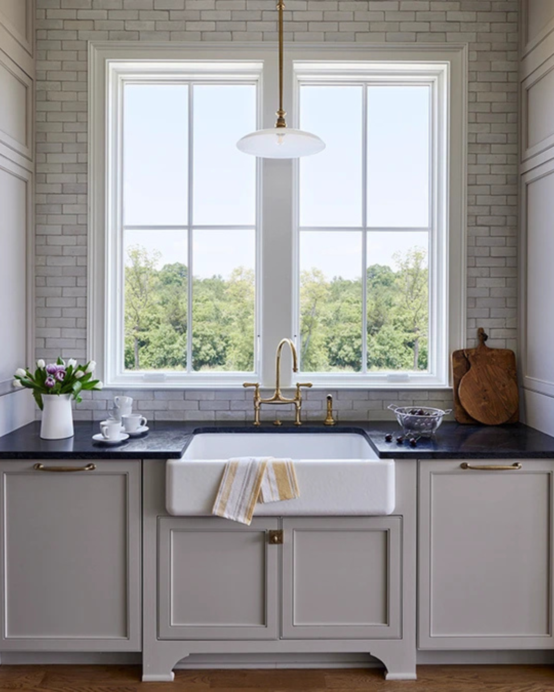 Kitchen with a large window, white brick walls, a farmhouse sink with a yellow and white towel, black countertop, and decorative items including a vase of flowers, white cups, a wooden cutting board, and a bowl of fruit.