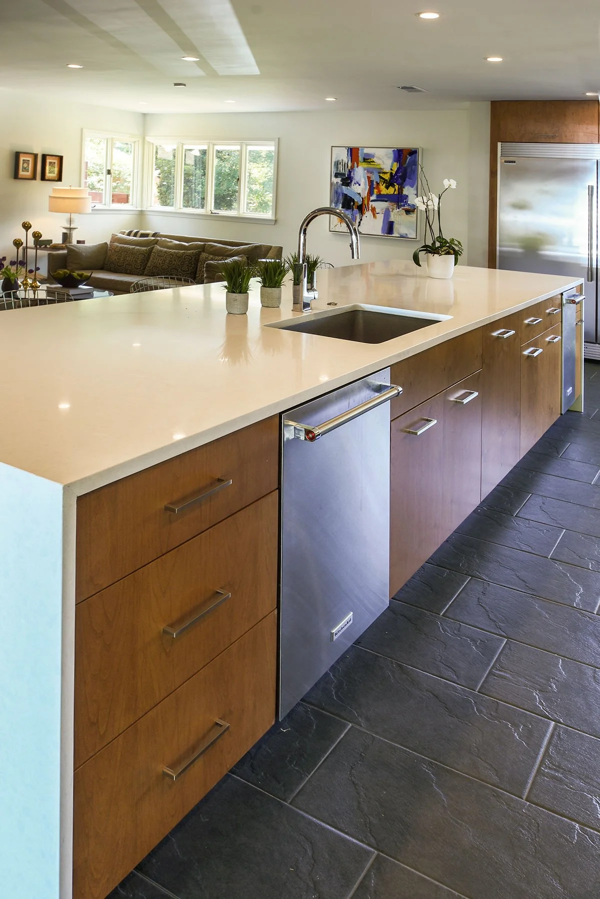 Open-concept kitchen with a large white countertop island, wooden cabinetry, black tiled flooring, and modern appliances. In the background, a living area with a brown sofa, colorful artwork, and plants.