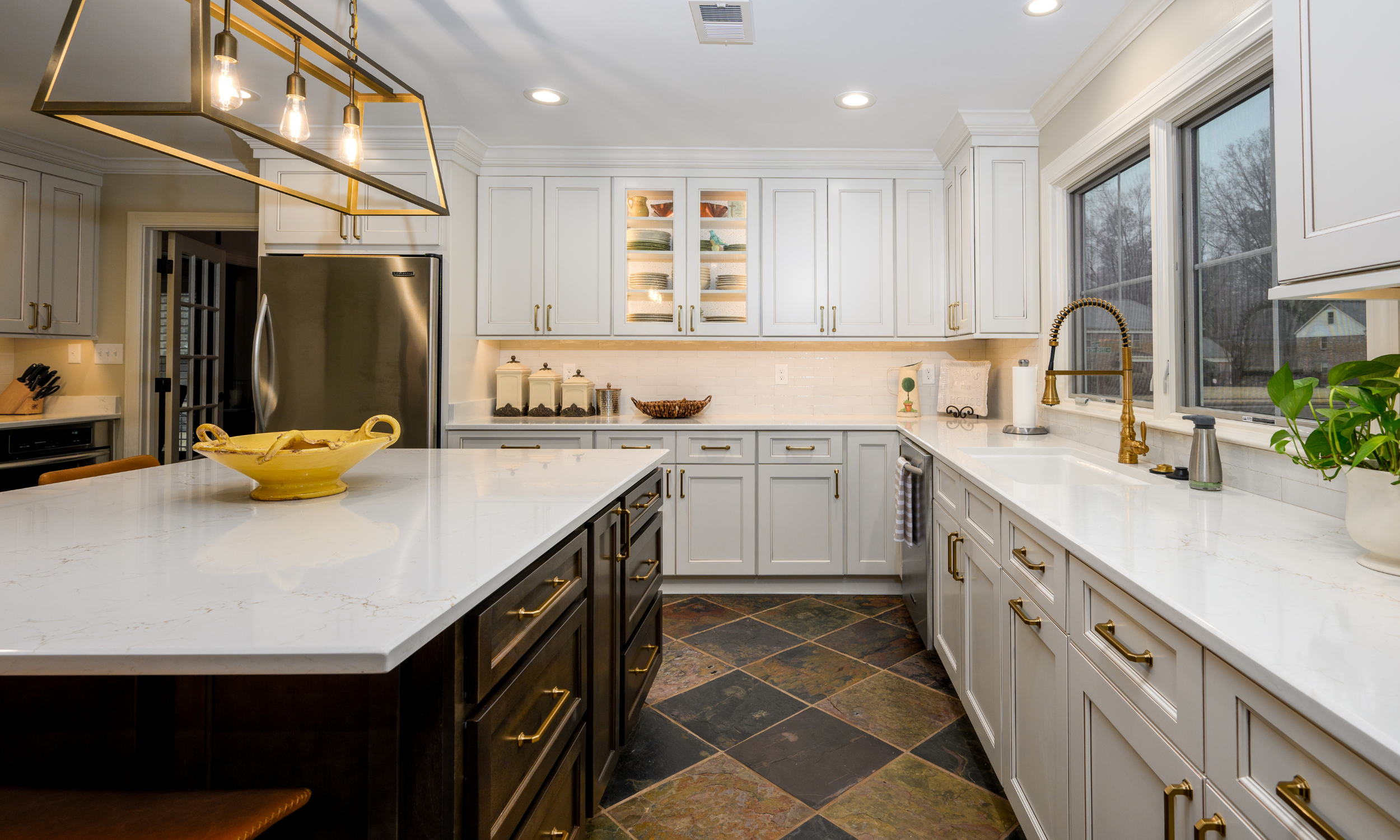 A modern kitchen with white cabinets, a large white marble island, and a window above the sink. There is a black and gold chandelier light fixture hanging above the island. The kitchen also features bronze hardware and a stainless steel refrigerator.